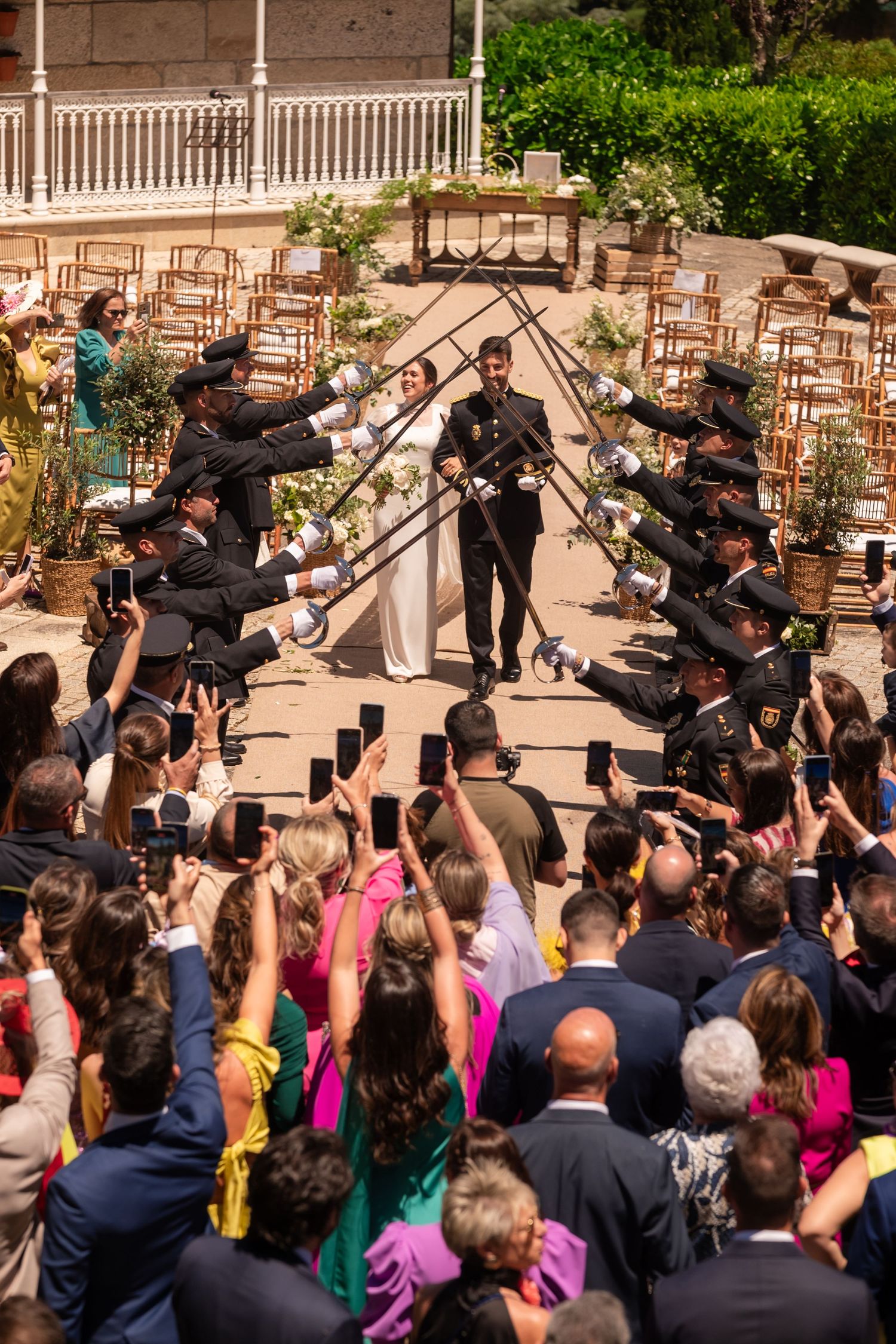 Military officers form sword arch as crowd gathers at outdoor wedding ceremony.