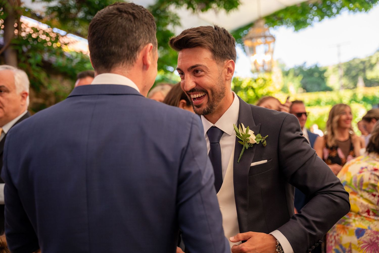 Wedding guests in formal attire gather outdoors for celebratory photos at a garden reception.