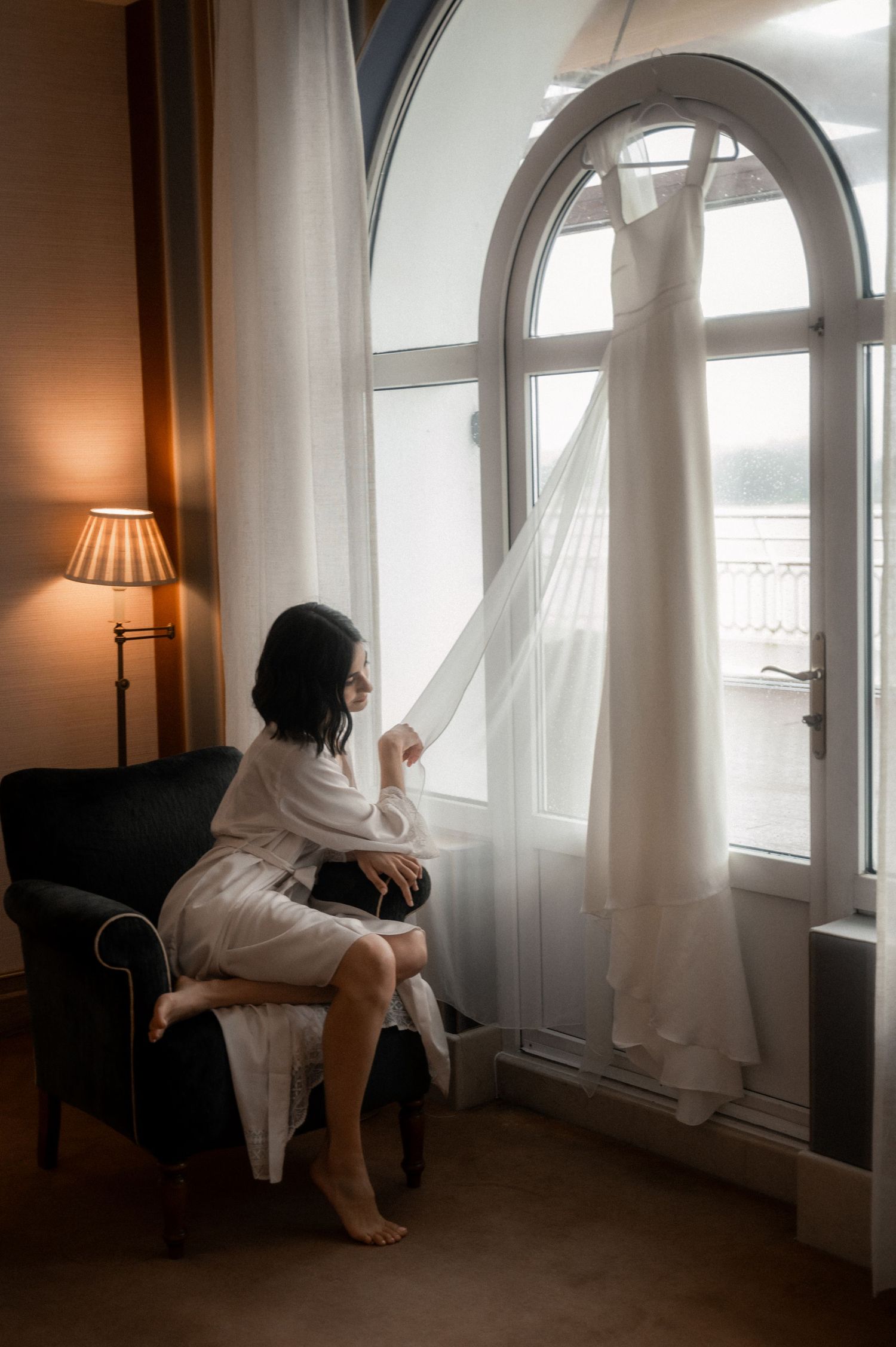 Moody artistic photos of a bride in a white dress sitting by large arched windows in warm natural light.