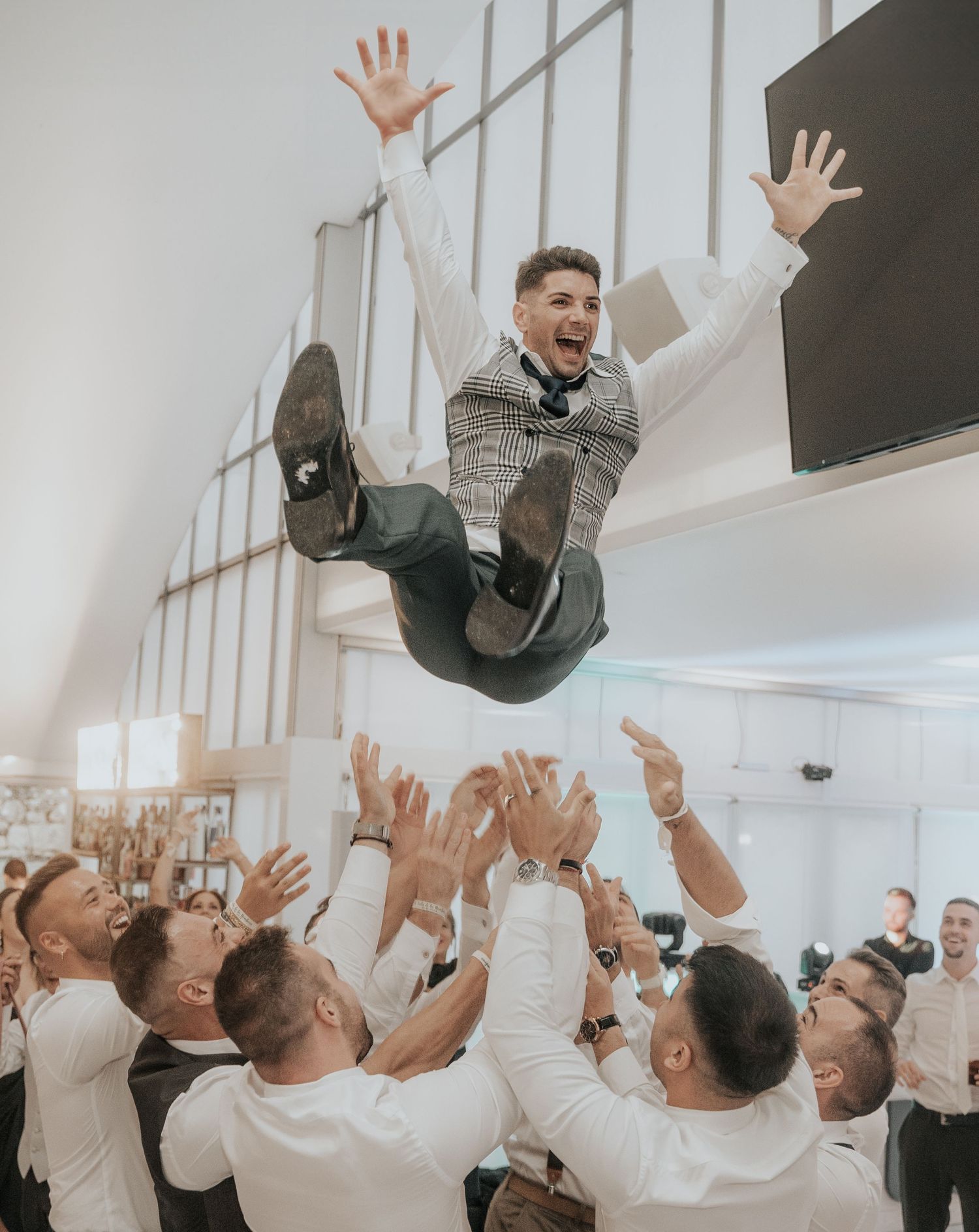 Groomsmen celebrating and tossing someone in the air during a joyful wedding celebration indoors.