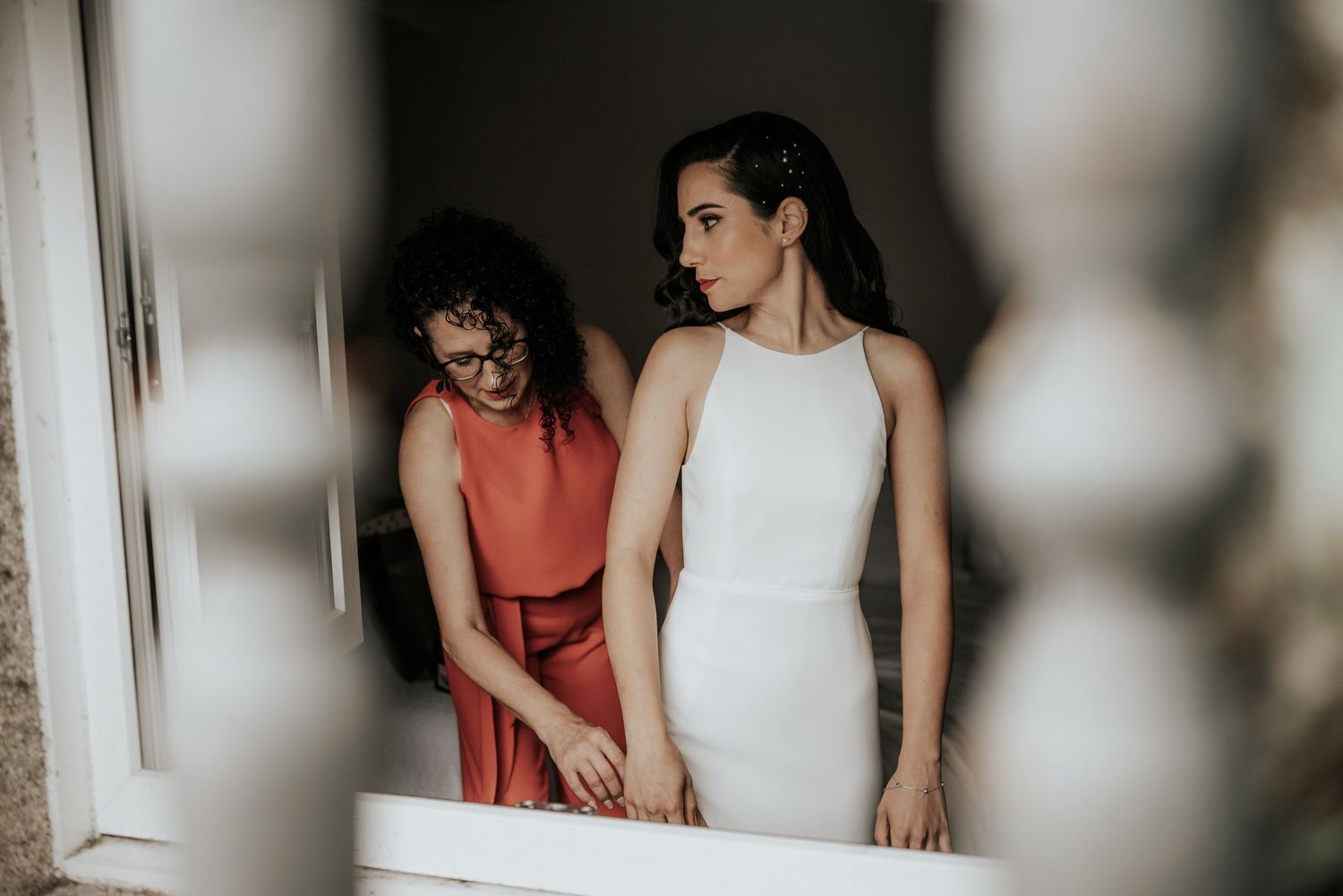 A couple in coral and white outfits pose together against a light-colored wall in a romantic photoshoot setting.
