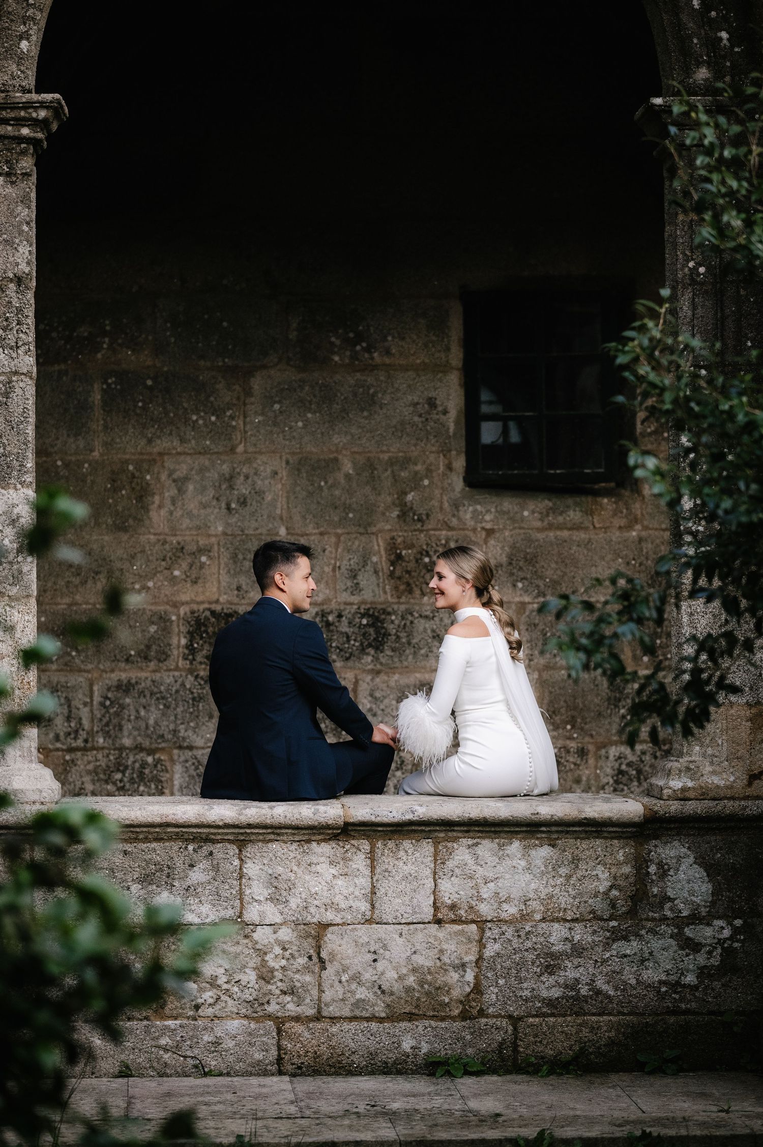 A couple in wedding attire sits together on a stone ledge in front of an old stone wall surrounded by greenery.