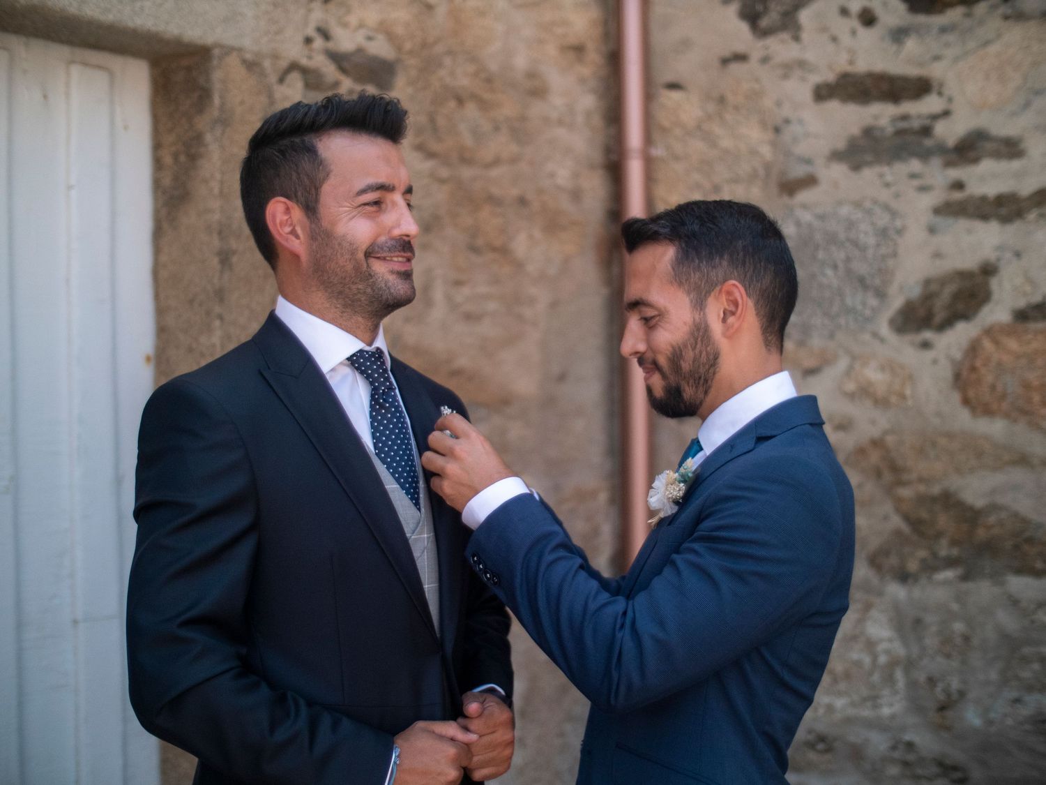 Two men in navy blue suits share a candid laughing moment against a stone wall background.