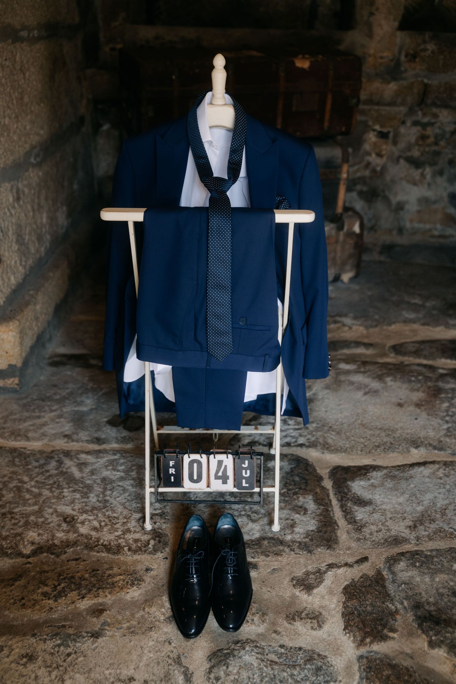Navy blue suit jacket and dress shoes arranged on white mannequin stand against rustic stone wall background.