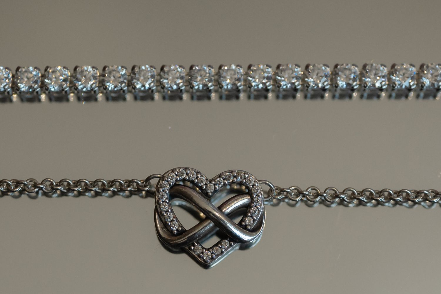Sparkling silver infinity heart pendant necklace with crystal accents alongside a tennis bracelet on beige background.