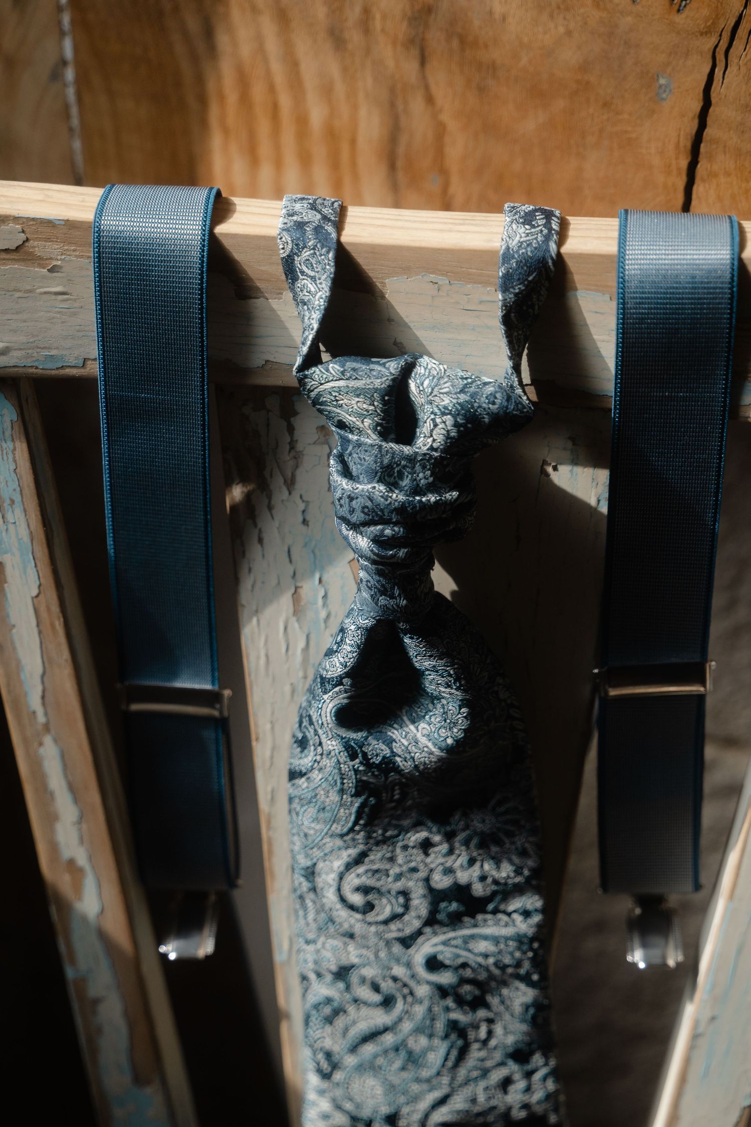 Navy blue suspenders and a matching patterned tie hanging on a wooden rack against a rustic background.