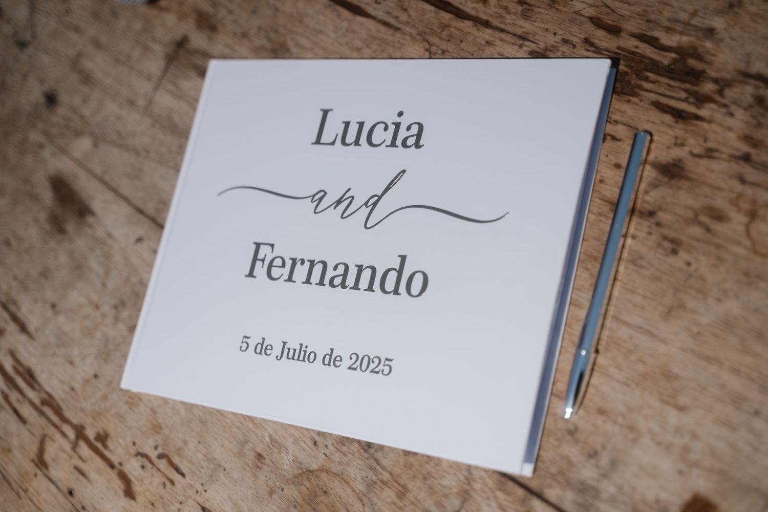 An elegant white wedding invitation card for Lucia and Fernando's July 2025 wedding rests on a wooden surface.