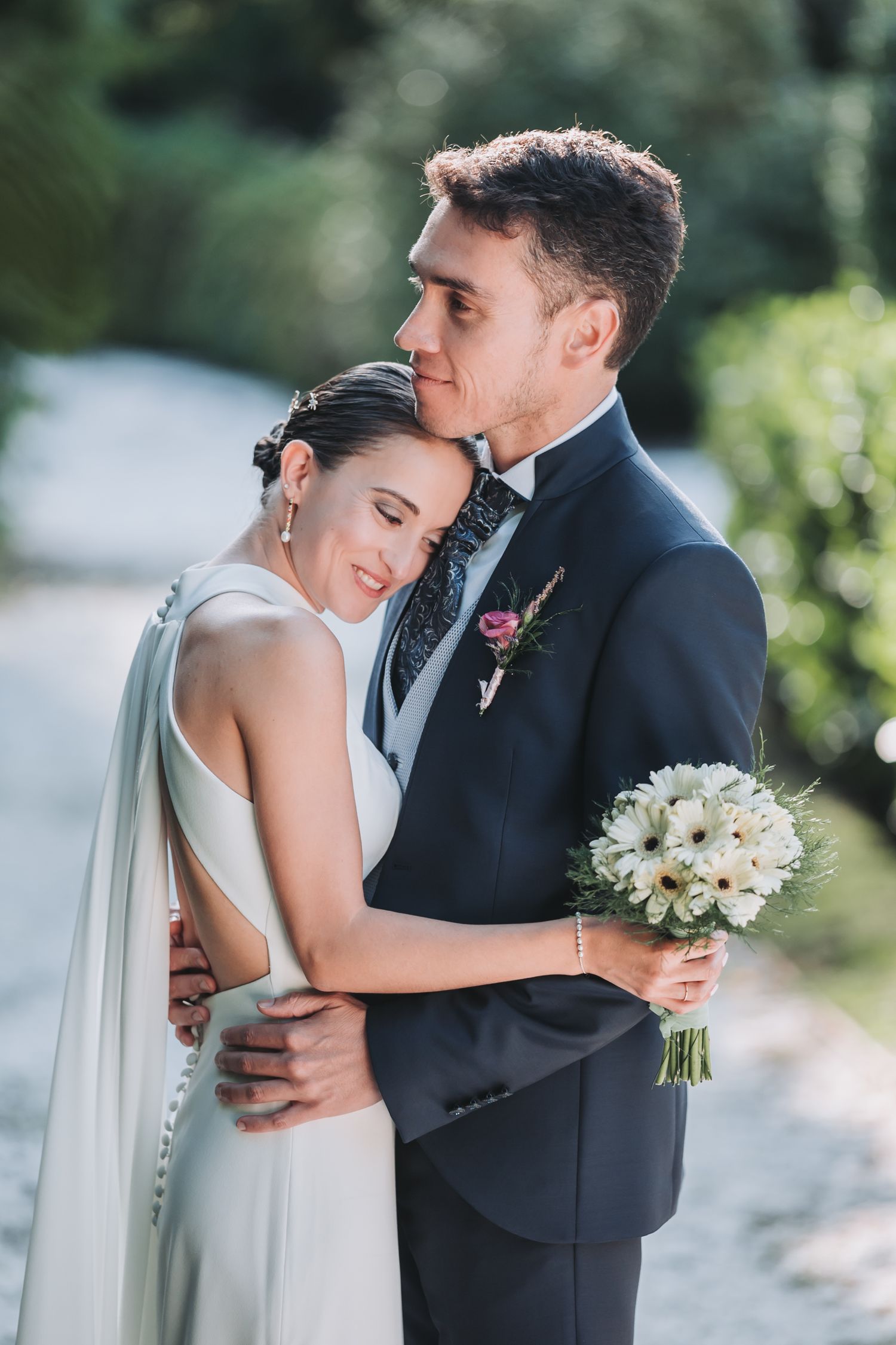 Romantic wedding couple embraces outdoors with bride holding white bouquet in elegant dress and groom in dark suit.