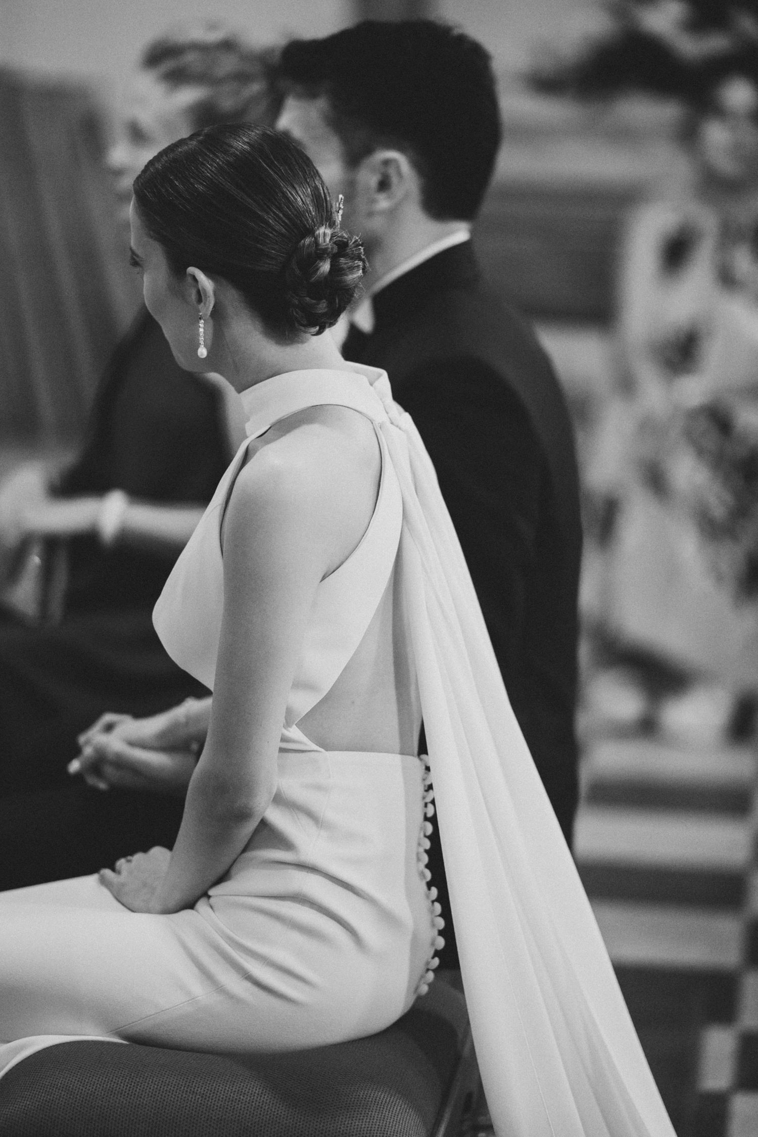 Black and white close-up photo of a bride in a sleeveless wedding dress with flowing veil sitting during ceremony.