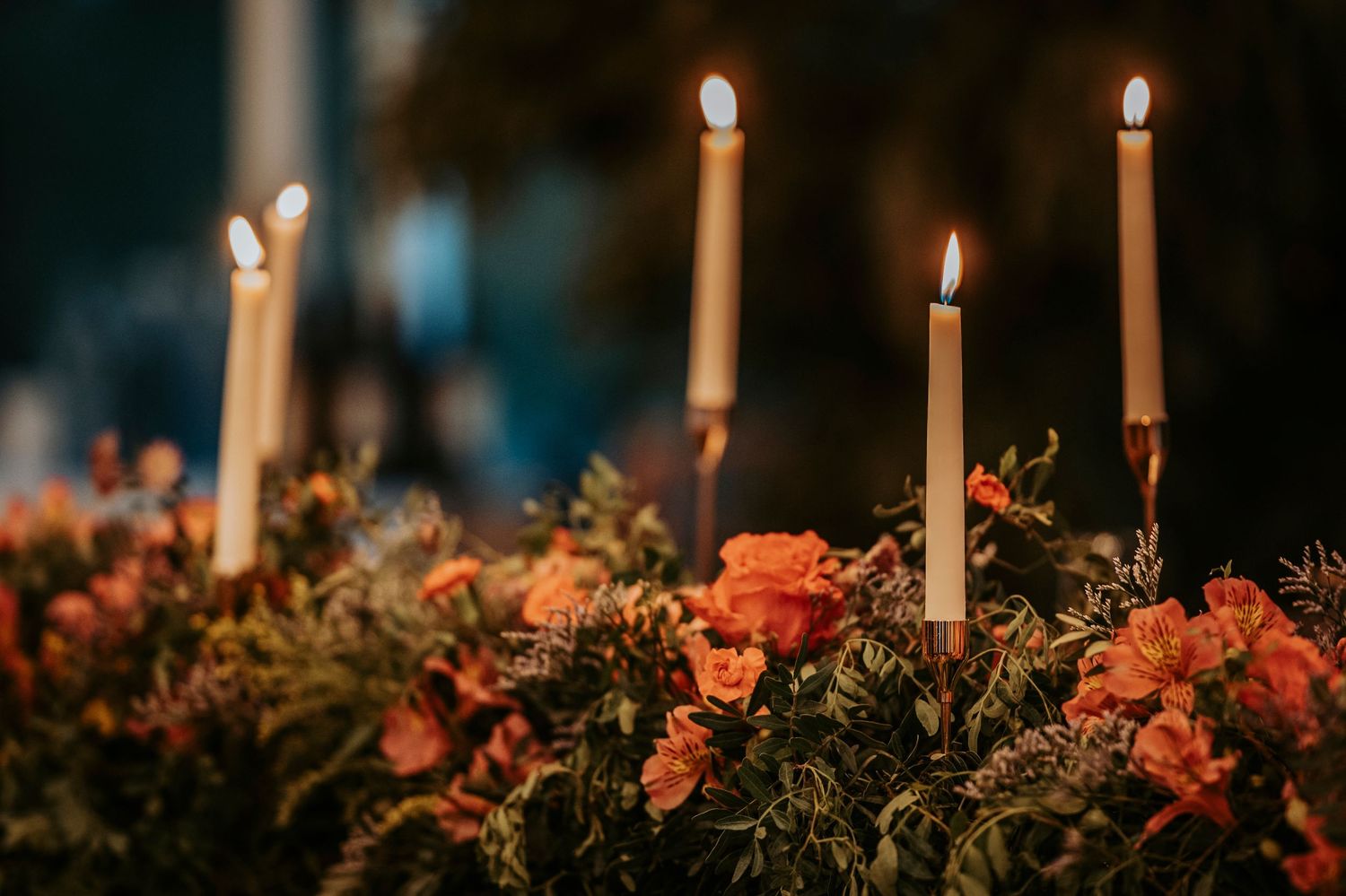 Glowing white candles illuminate floral arrangements of coral roses and greenery in a romantic tablescape setting.