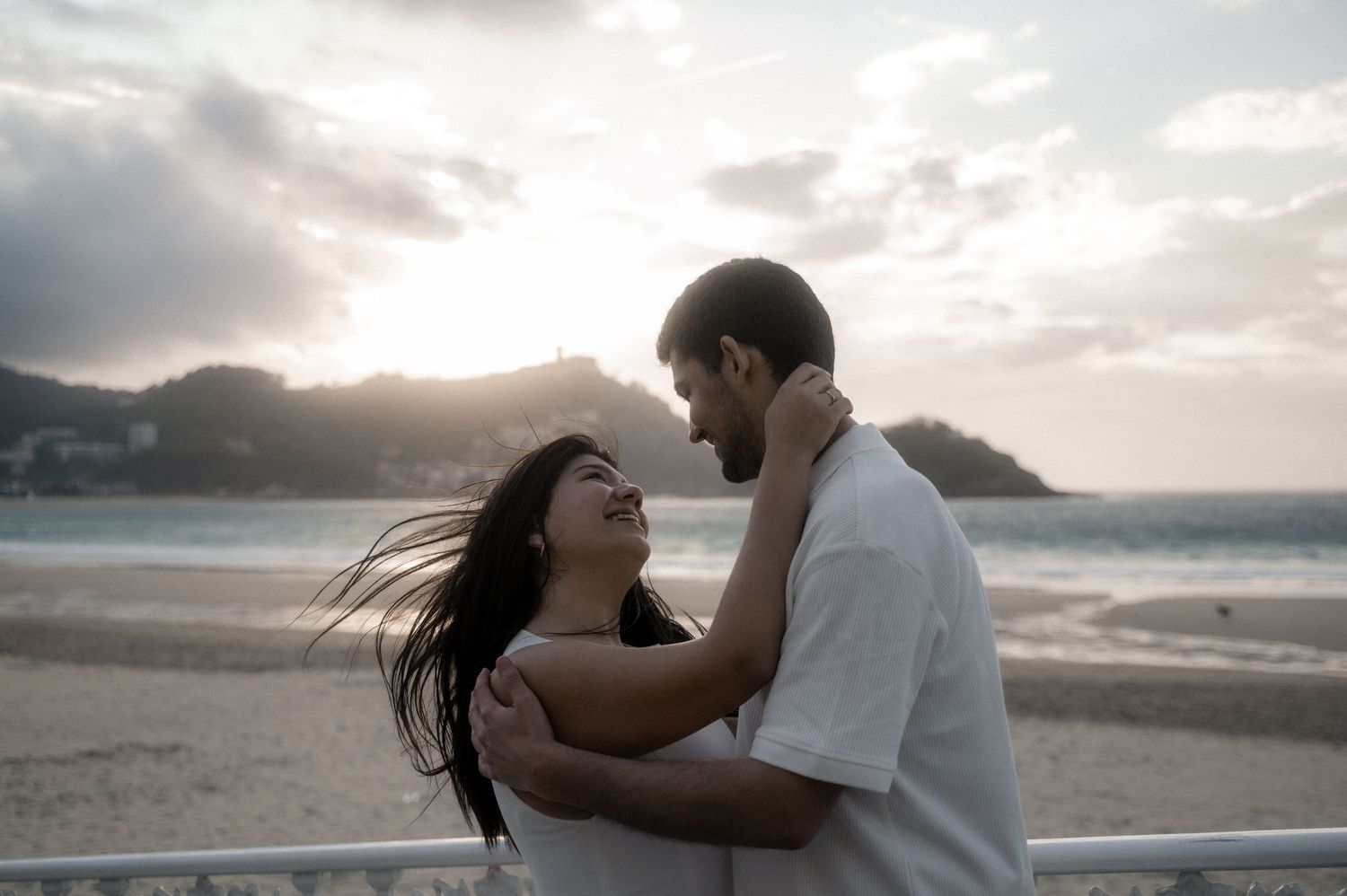 A couple shares an intimate moment on a beach at sunset with waves and cloudy skies in the background.