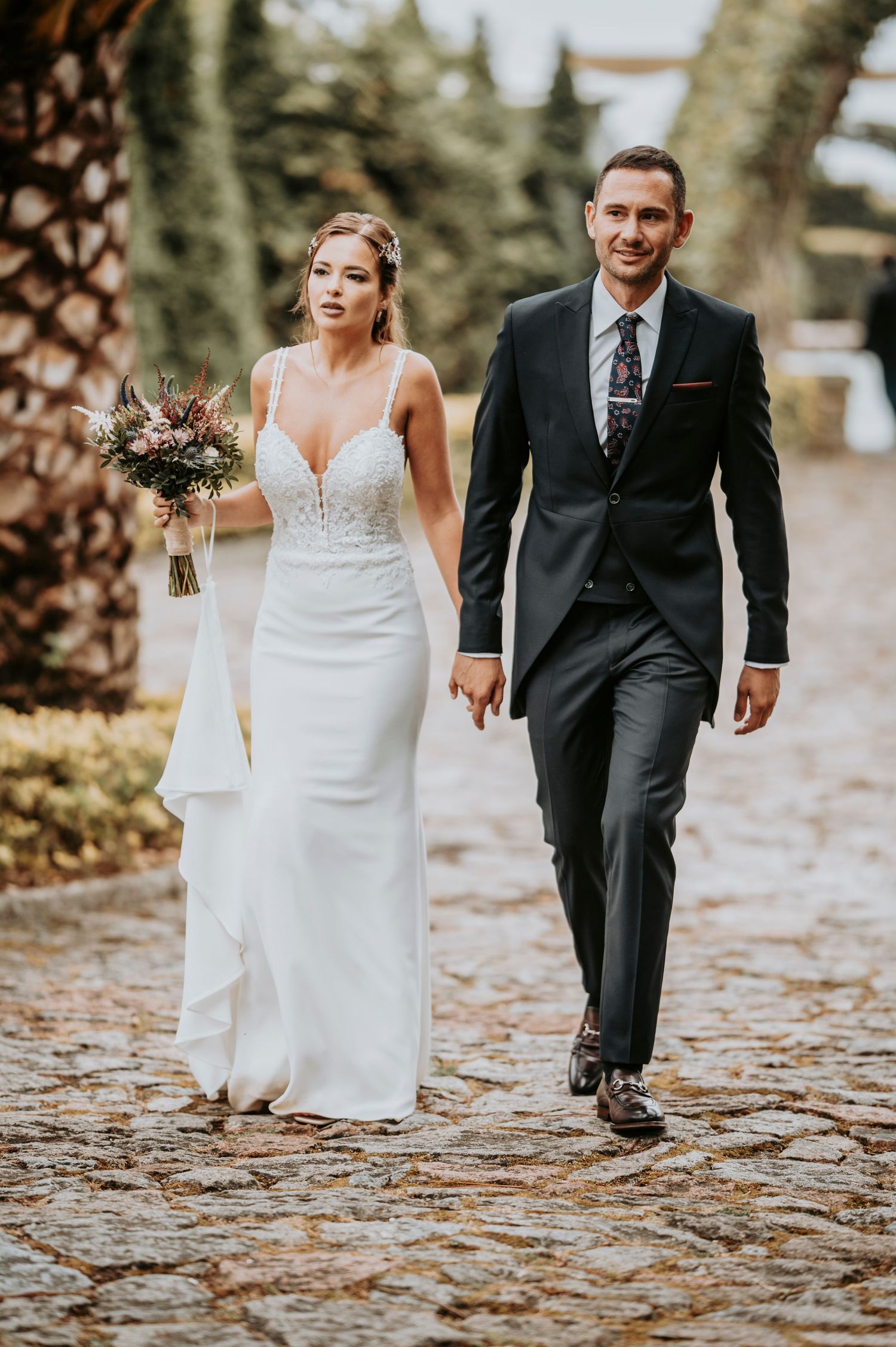 A couple walking hand in hand on a path with fallen leaves, the bride in a white dress and groom in a black suit.
