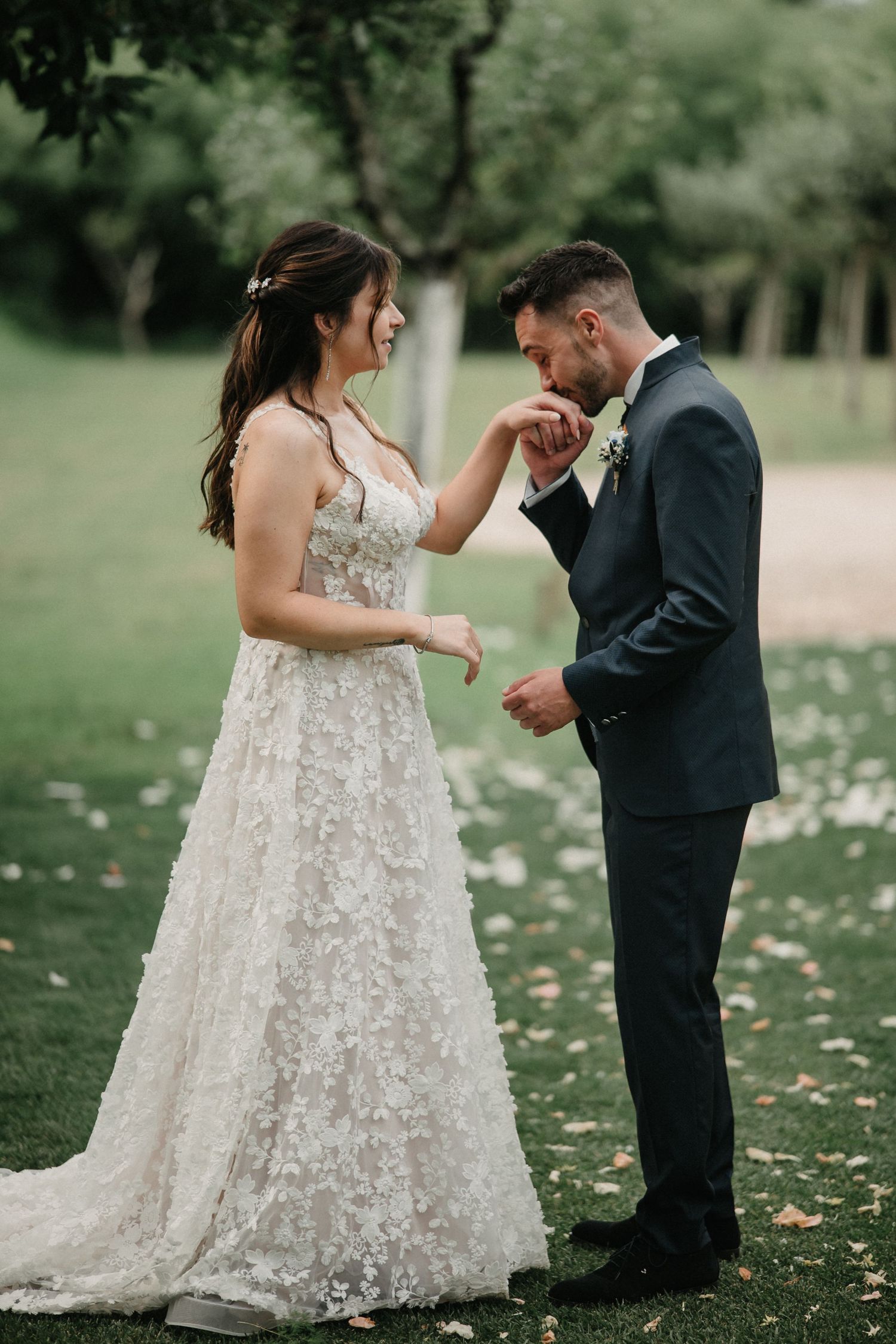 A romantic wedding moment captured in a lush green outdoor setting with a bride in a floral lace gown and groom in black suit.