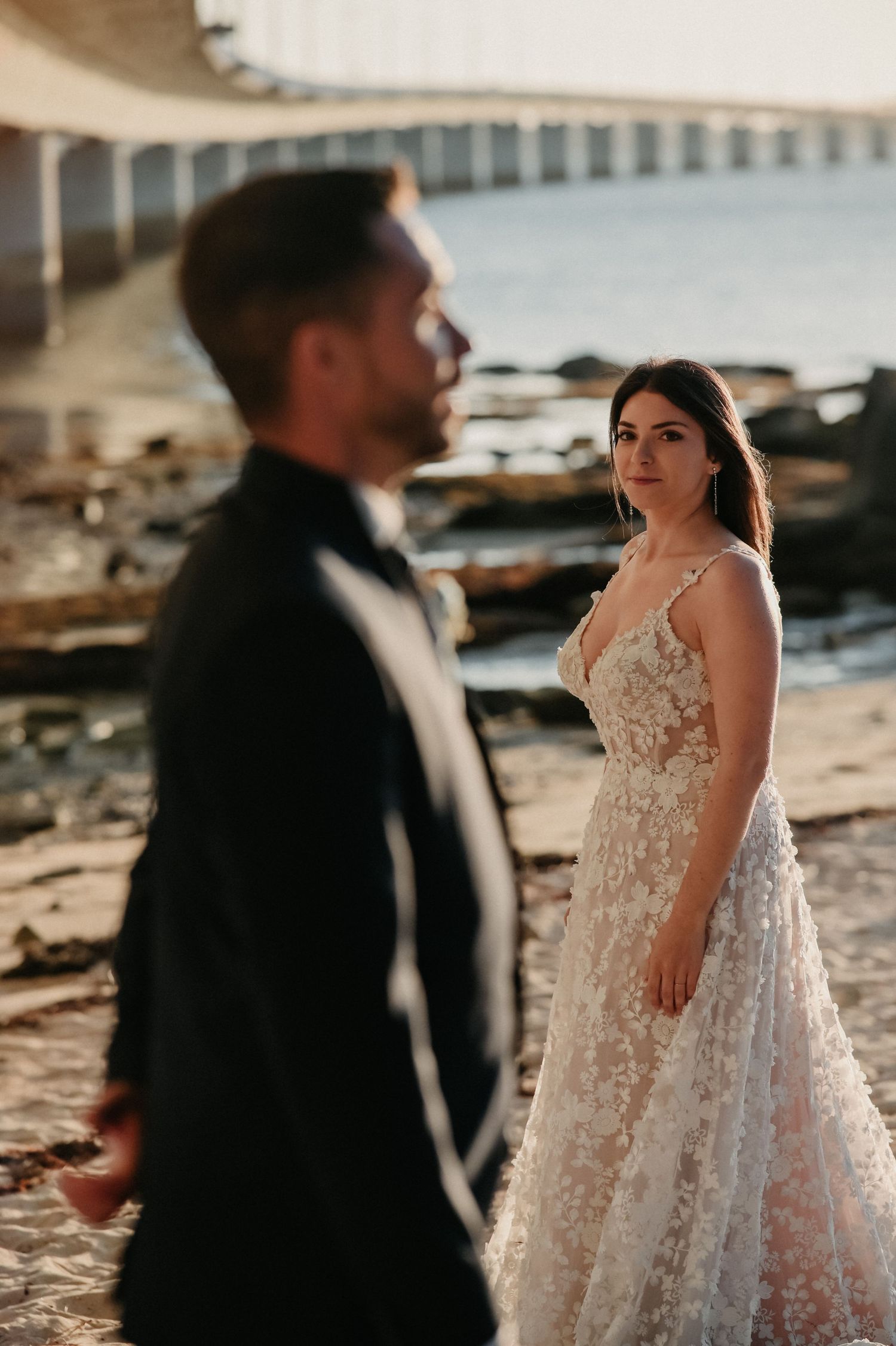 Newlywed couple in formal attire poses on a rocky beach during golden hour.