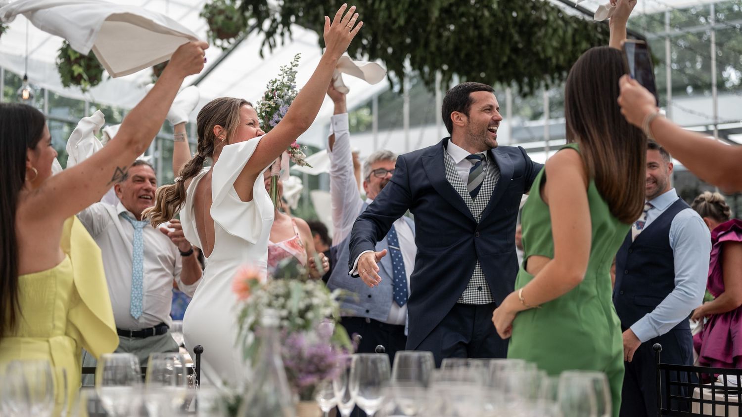 Wedding guests dancing and celebrating with raised arms at an outdoor reception under string lights and trees.