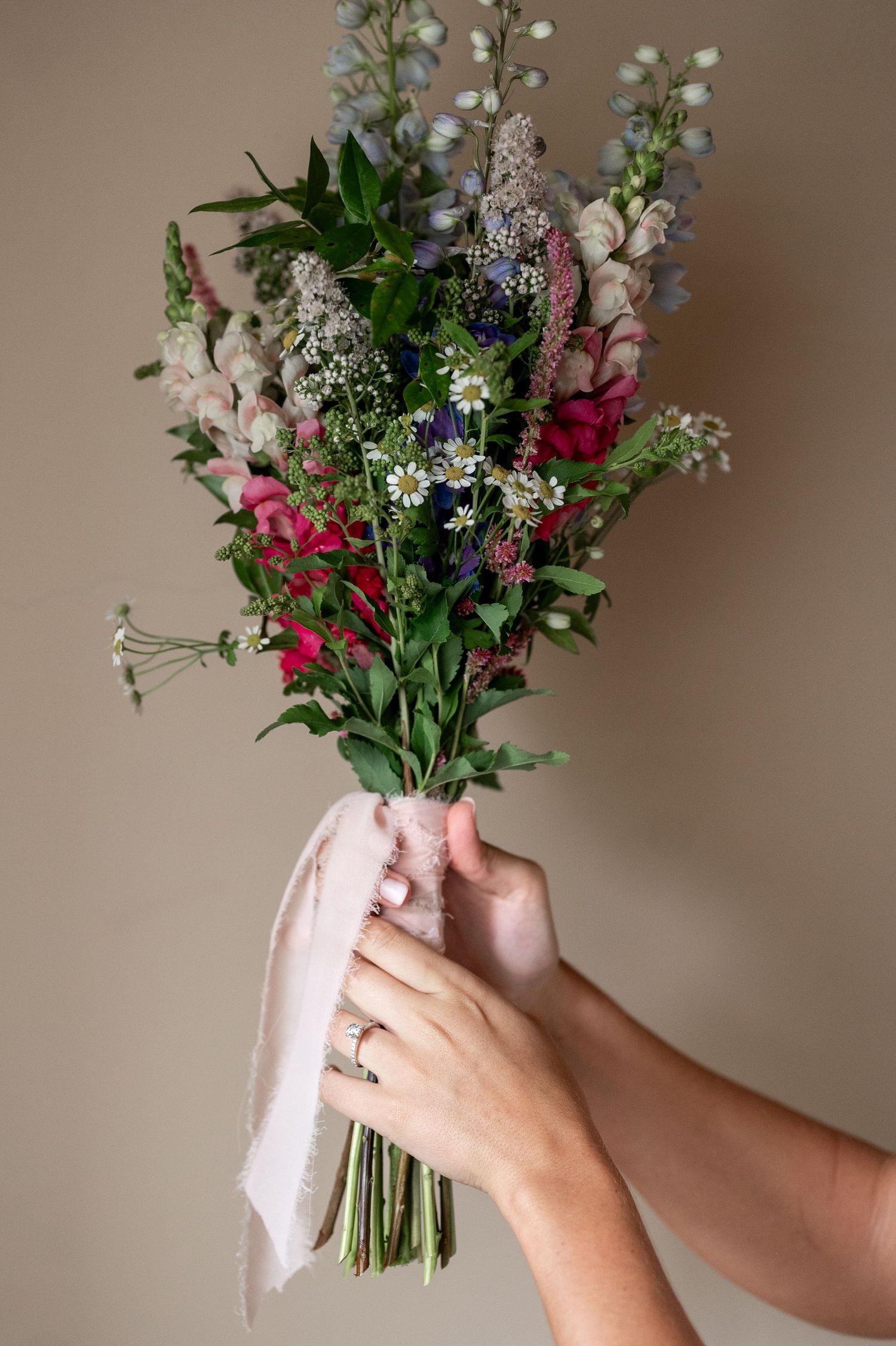 Hands holding a fresh wildflower bouquet with pink, purple, and white blooms wrapped in white fabric.