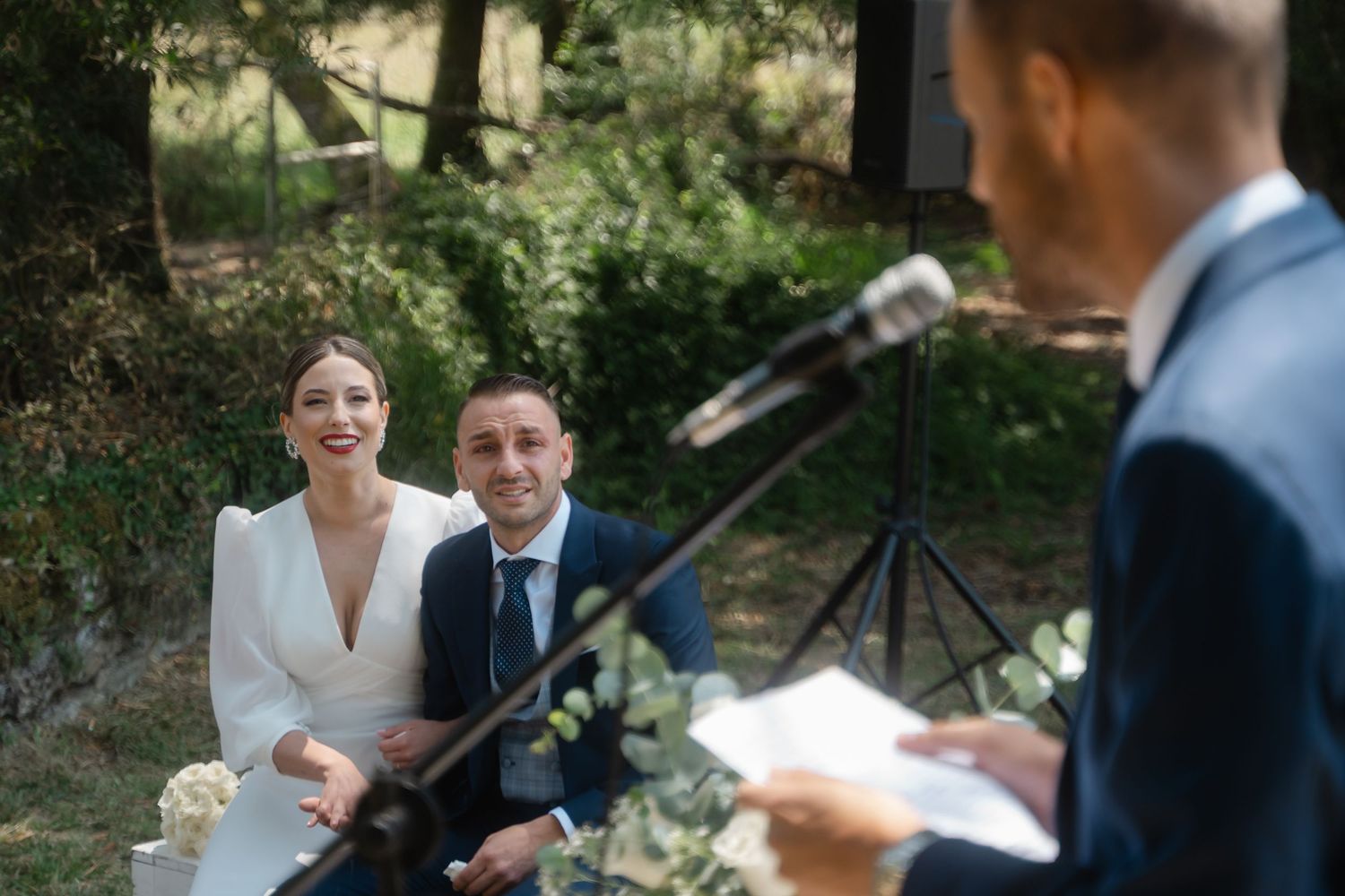 A couple shares a joyful moment during their outdoor wedding ceremony in a sunlit garden setting.