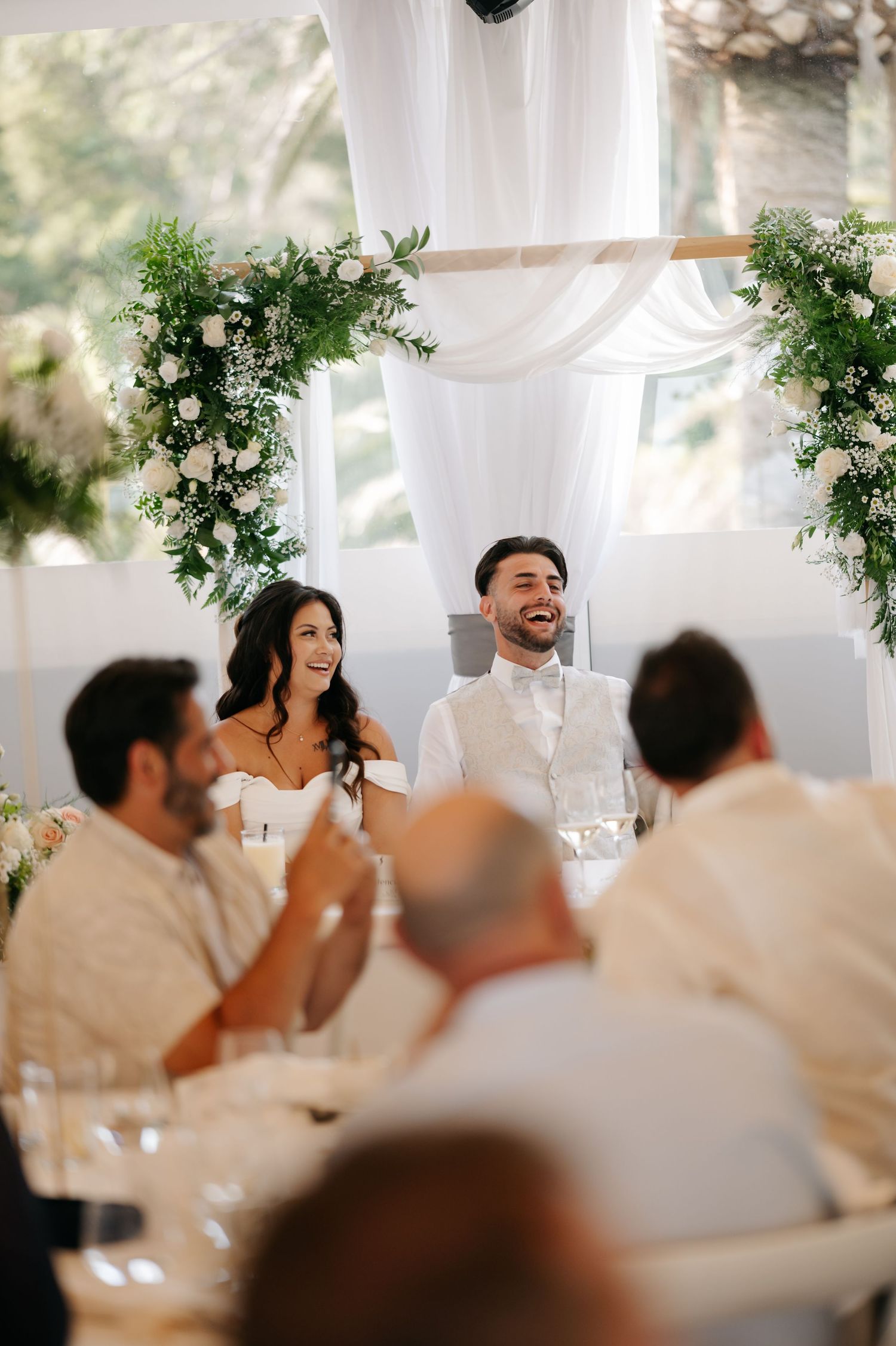 Wedding reception guests seated at tables with white decor and greenery arrangements in an elegant venue.