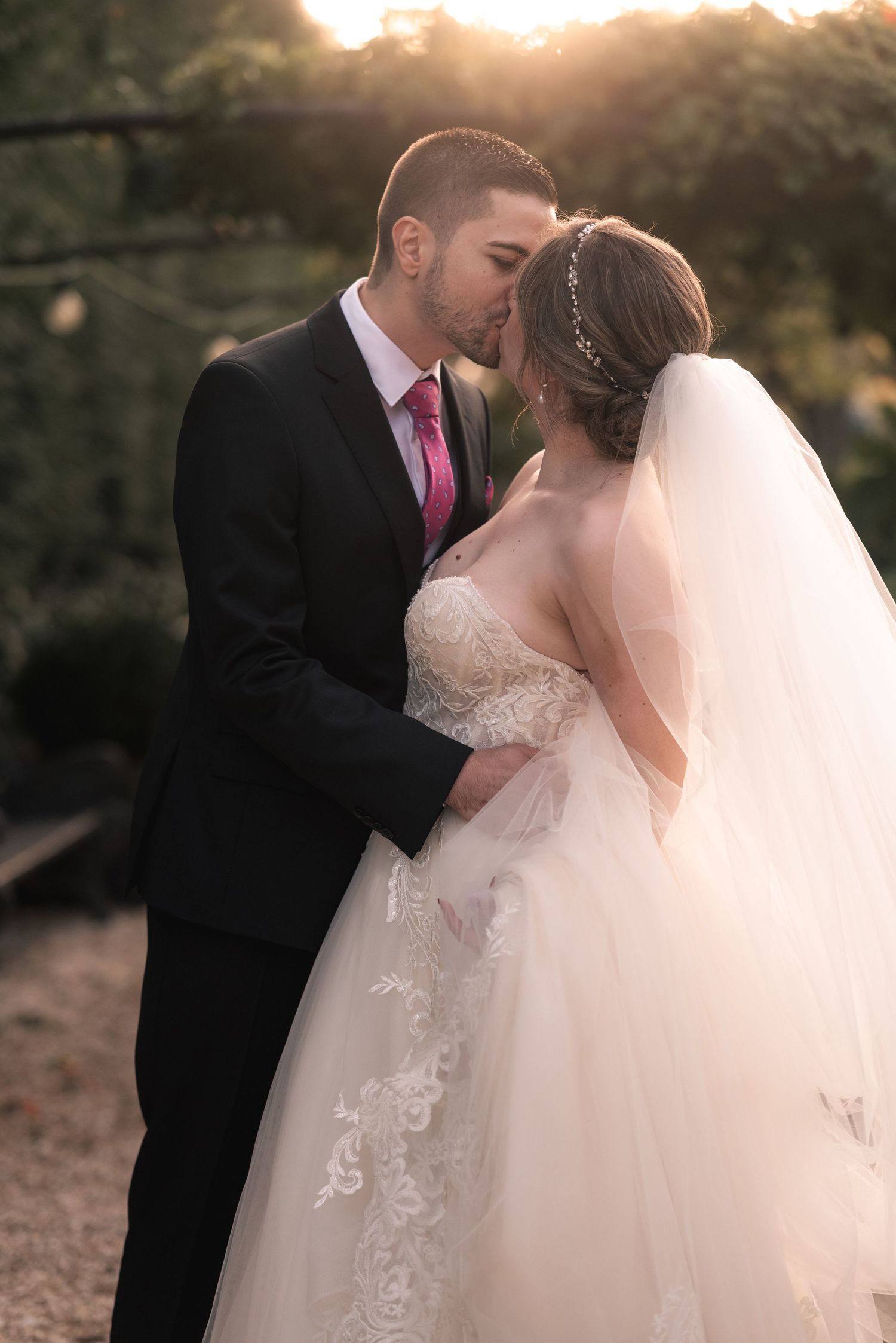 A romantic sunset wedding photo of a couple embracing, with the bride in a lace gown and the groom in a black suit and pink tie.