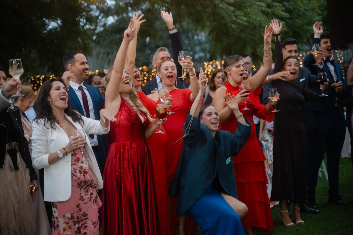 Wedding guests in colorful attire raise their hands in celebration during an outdoor reception.