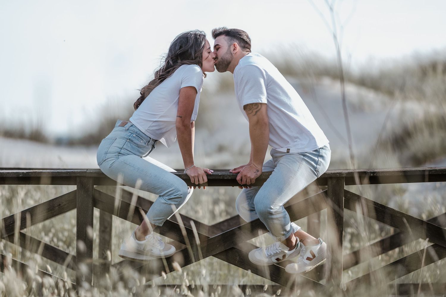 Two people in white clothing sitting on a wooden fence in a grassy field.