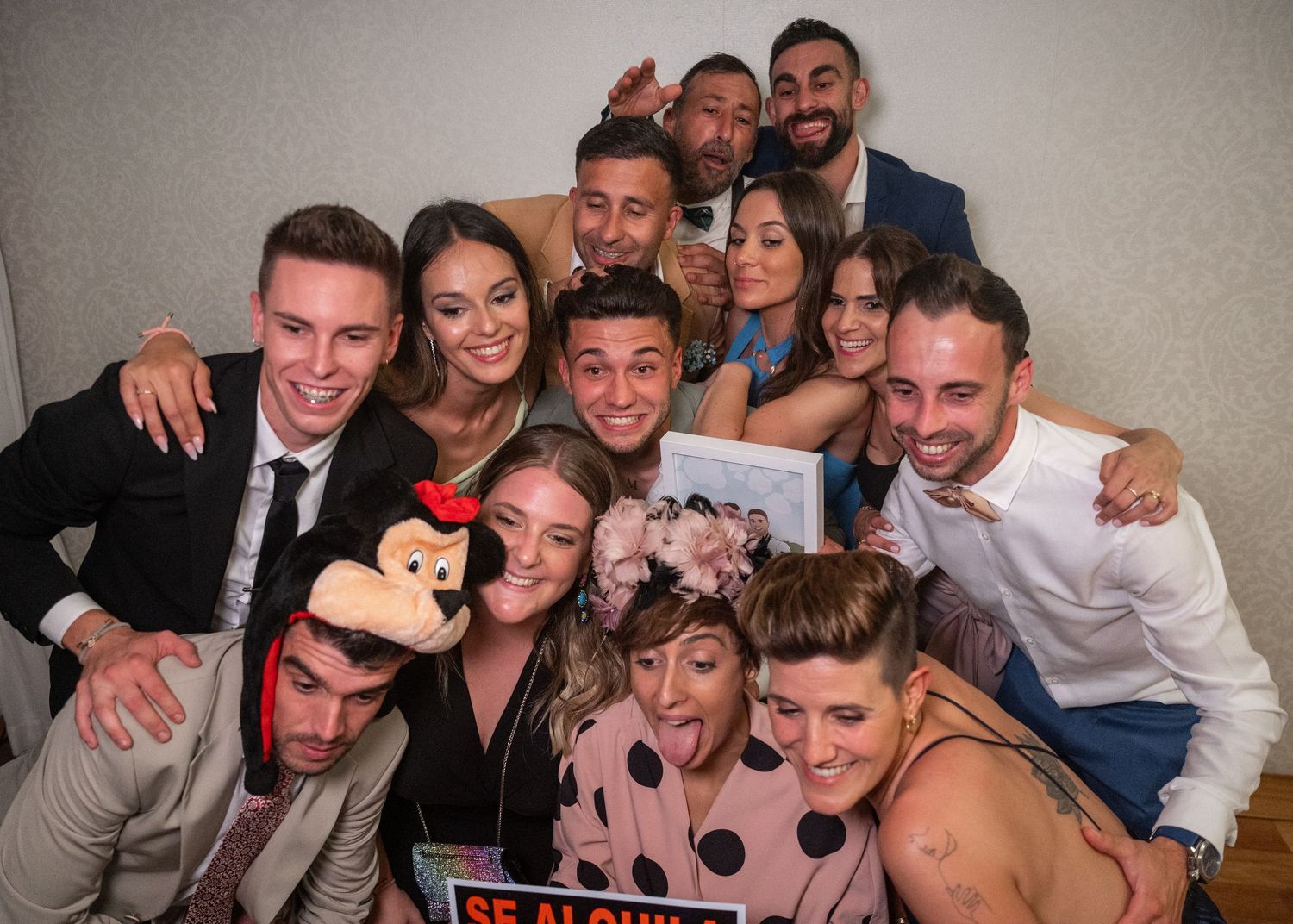A group of friends pose together at a party with a Mickey Mouse prop and pink floral decorations.