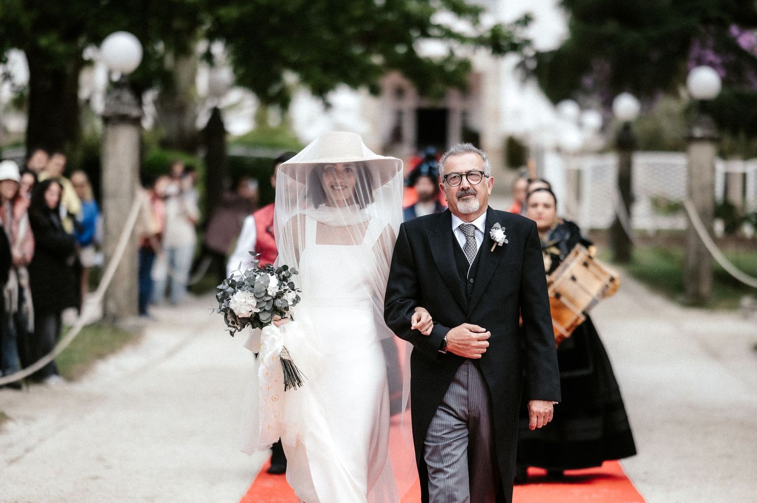 A wedding procession walking along a stone path surrounded by trees and guests.