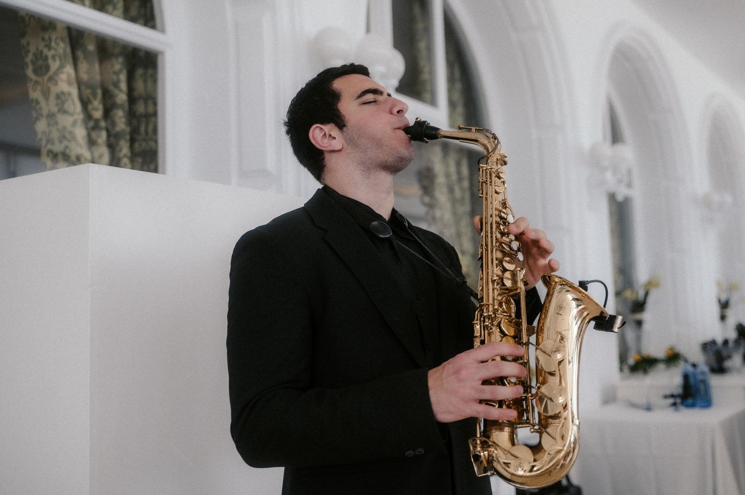 A musician plays a saxophone while standing against a white wall in a performance setting.