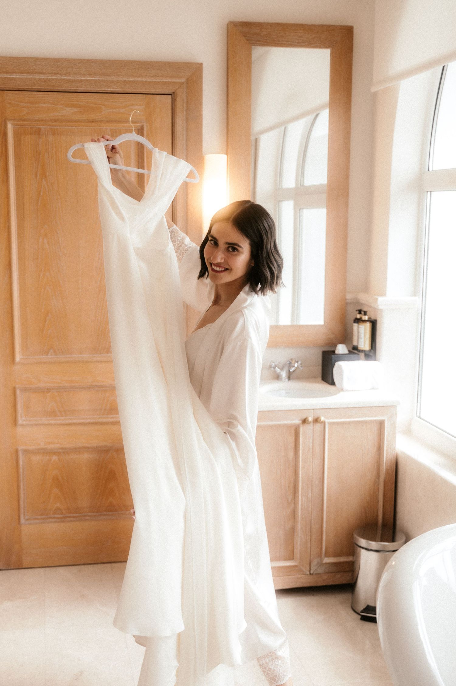 A series of ethereal white wedding dresses hanging on hangers in a sunlit dressing room with wooden paneled walls.