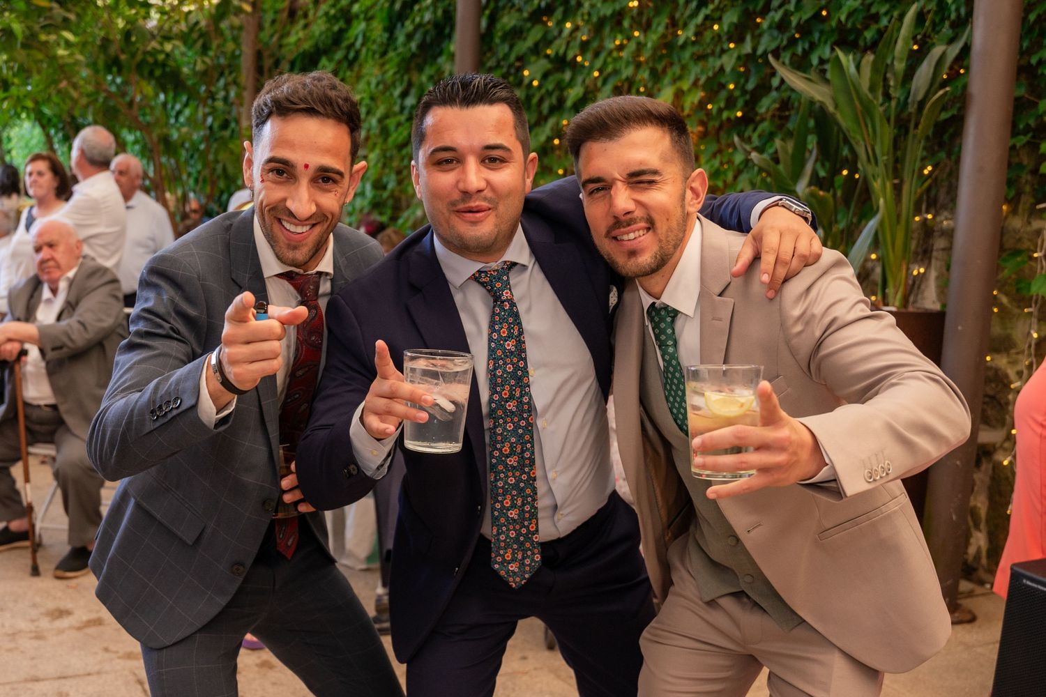 Groomsmen in navy suits pose together with thumbs up gestures at an outdoor celebration.