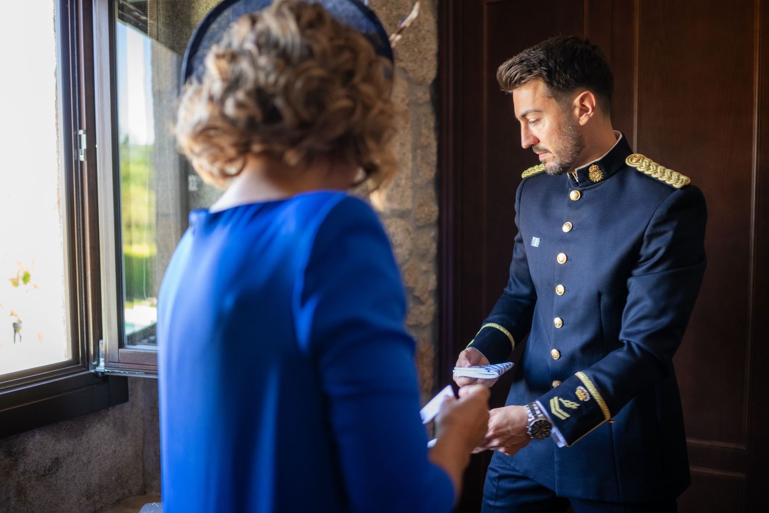 A couple in formal attire stands near a window, with one person wearing a bright blue dress and another in a naval uniform.