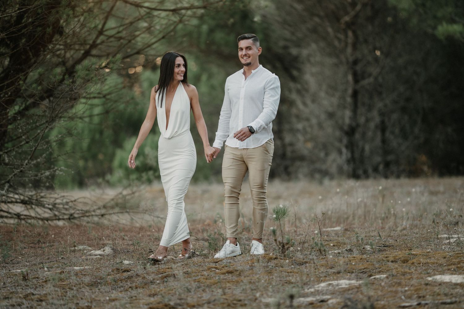 A couple in white and beige attire walking together through a wooded outdoor setting during a photoshoot session.