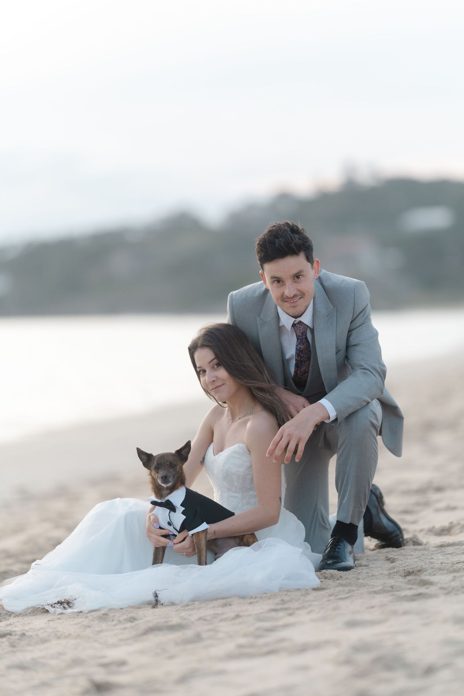 Bride and groom pose with small dog in tuxedo on sandy beach at dusk during romantic wedding photo shoot.