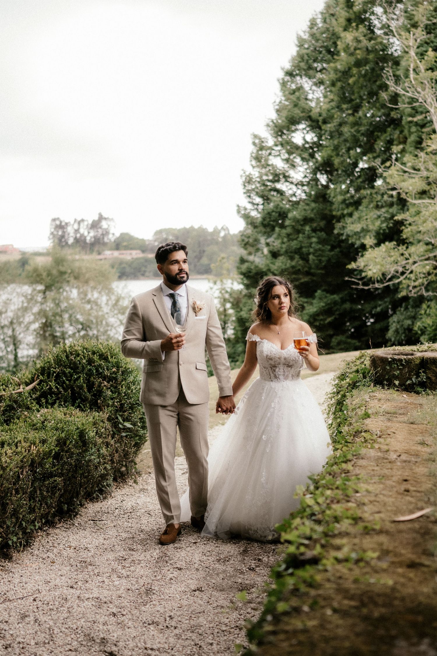 A couple walks hand in hand along a garden path, the bride in a white gown and groom in a light beige suit.