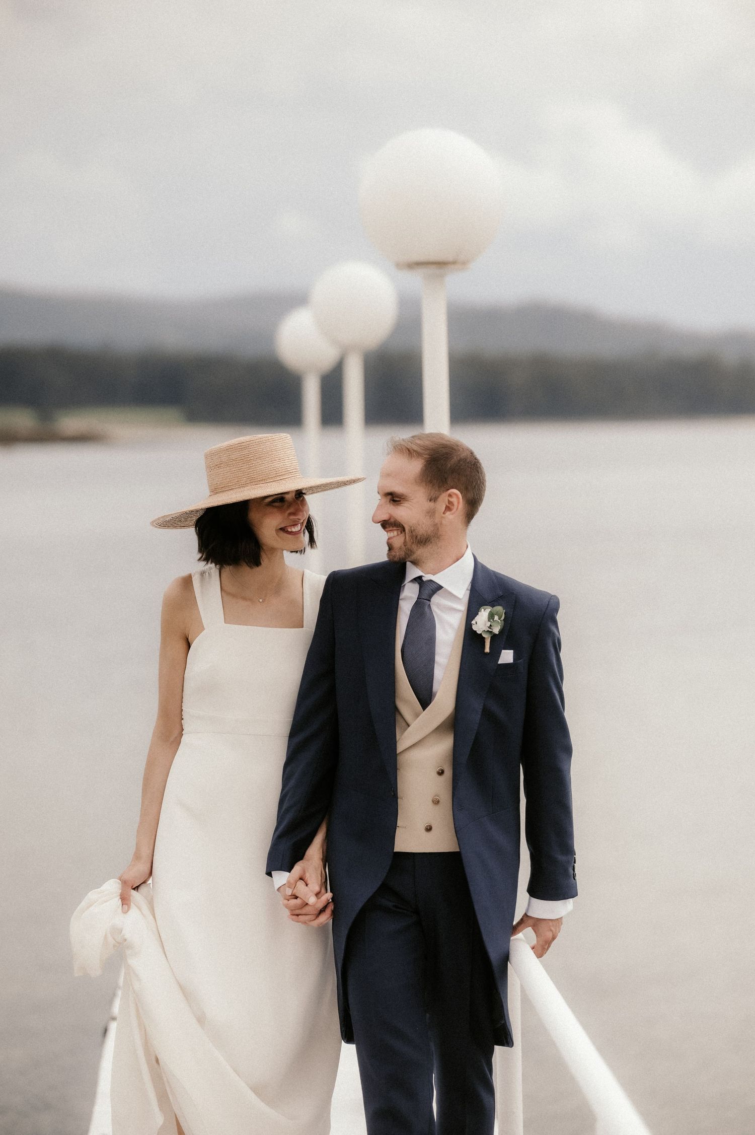 Elegant wedding couple poses by a serene lake with mountains in background and bride in wide-brimmed hat.