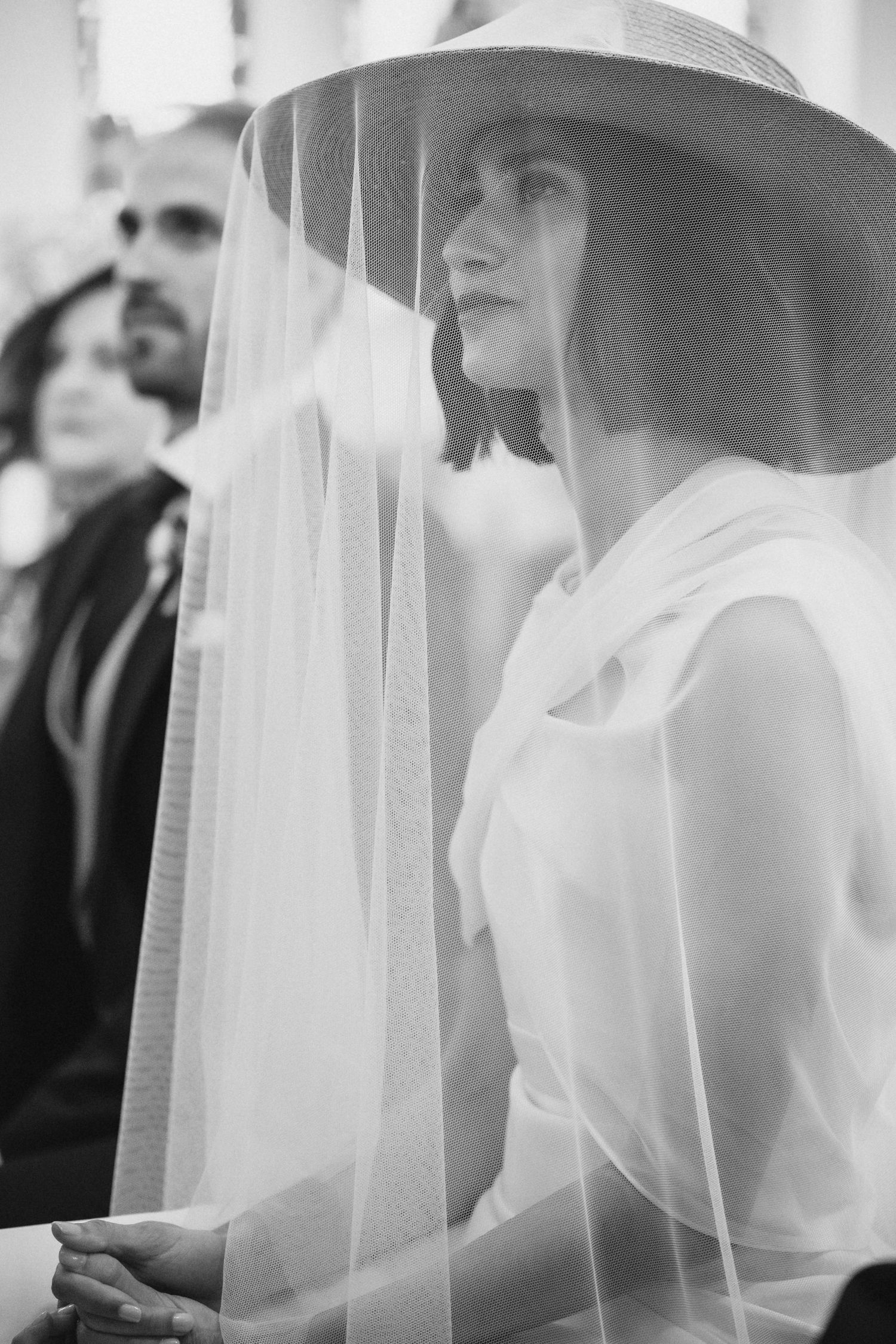 Black and white photo of a bride in a minimalist dress and dramatic wide-brimmed hat veil during ceremony.