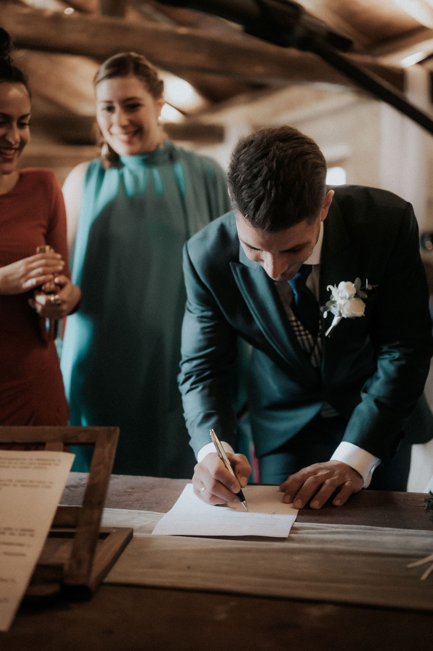 Wedding guests in formal attire sign documents at a wooden table during the ceremony.