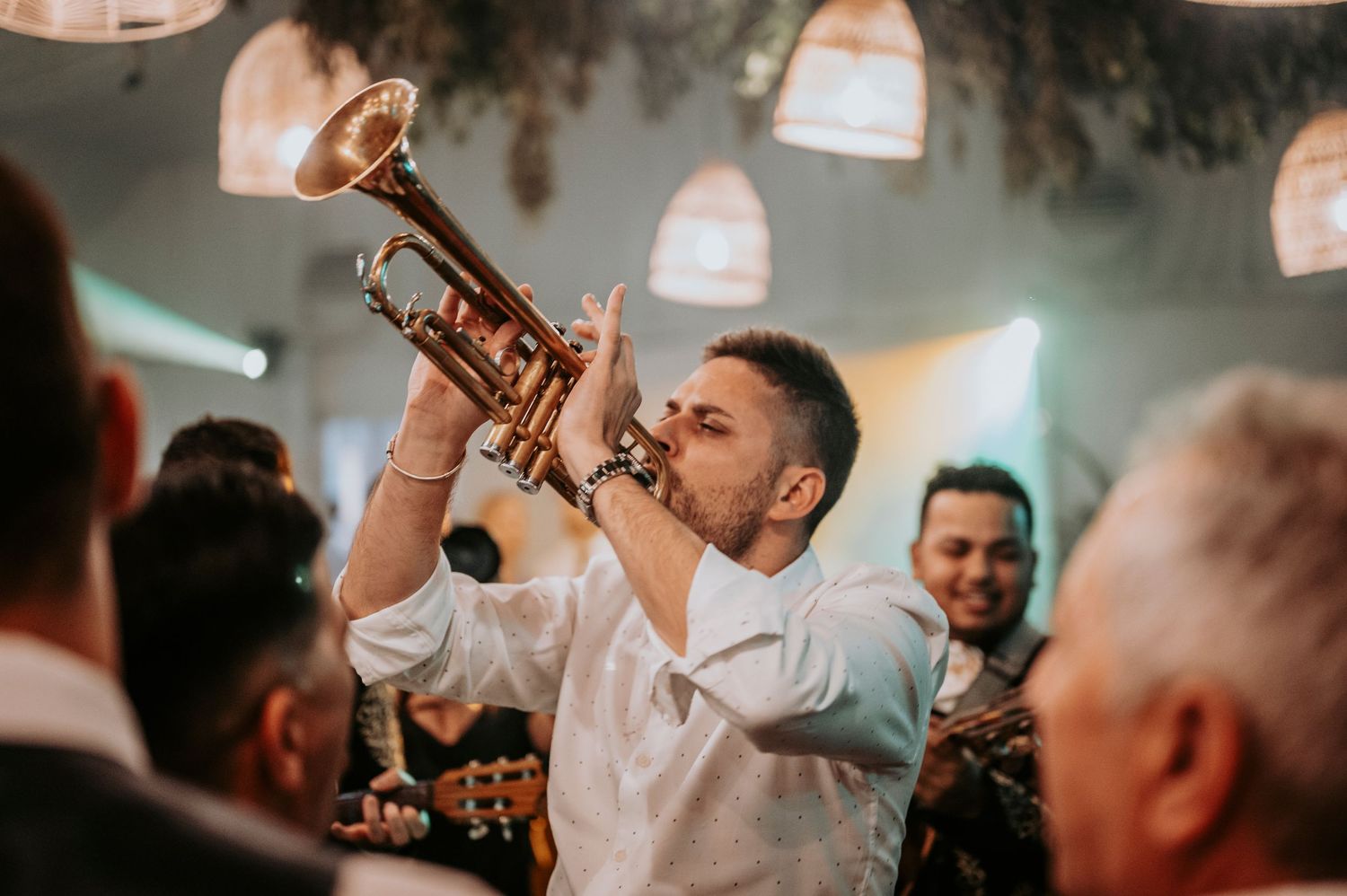 A trumpet player performs with enthusiasm at an indoor venue with hanging pendant lights and a festive atmosphere.