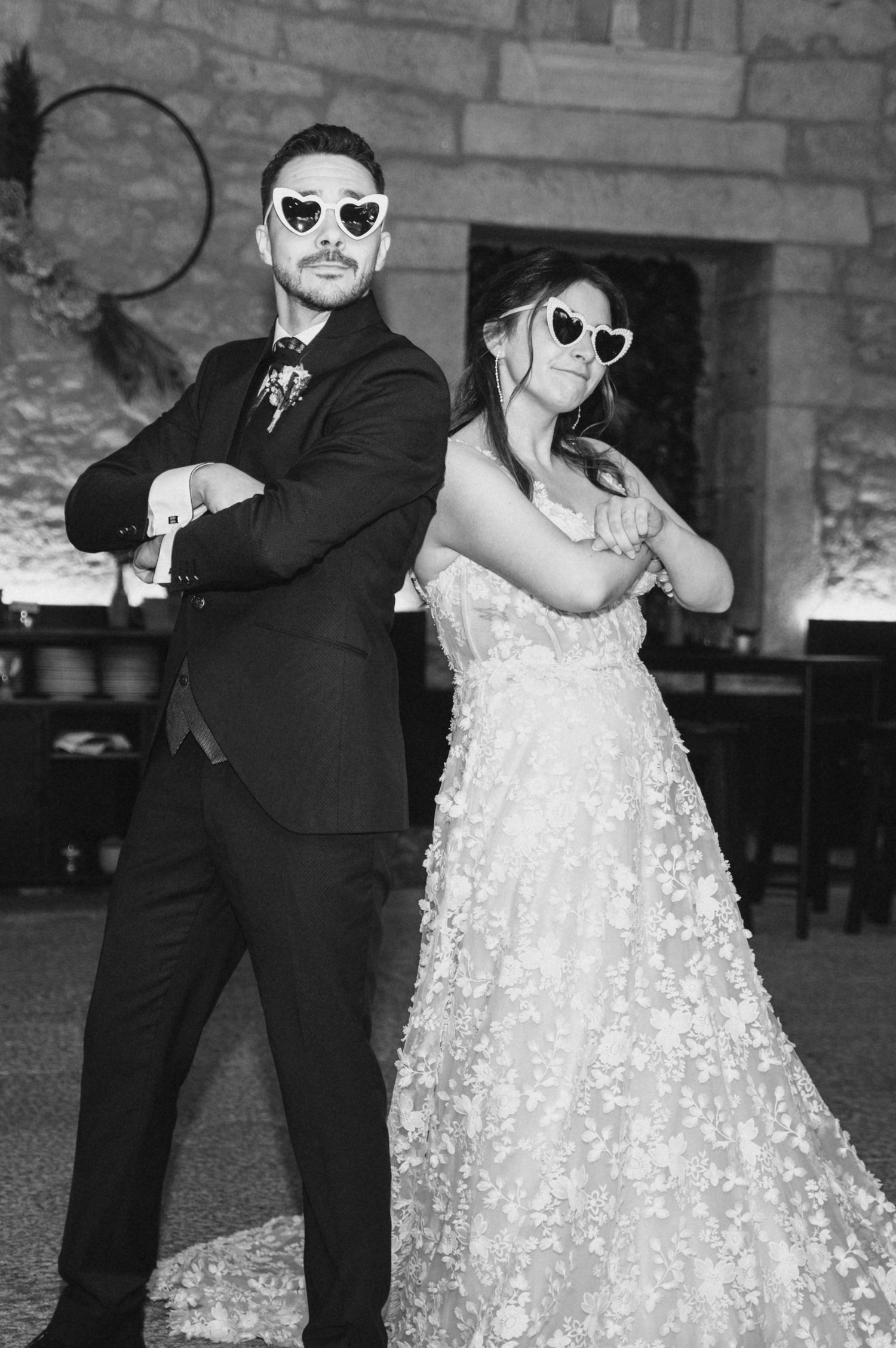 Stylish couple in sunglasses strike fun poses, with bride wearing sparkly gown and groom in classic black suit.