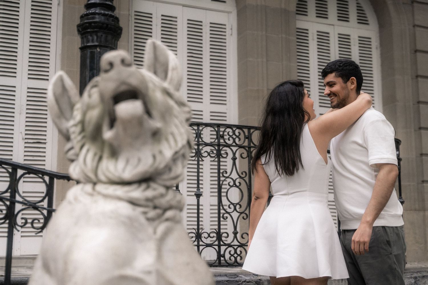 A romantic scene unfolds on an elegant balcony with white shutters and classical architecture in the background.