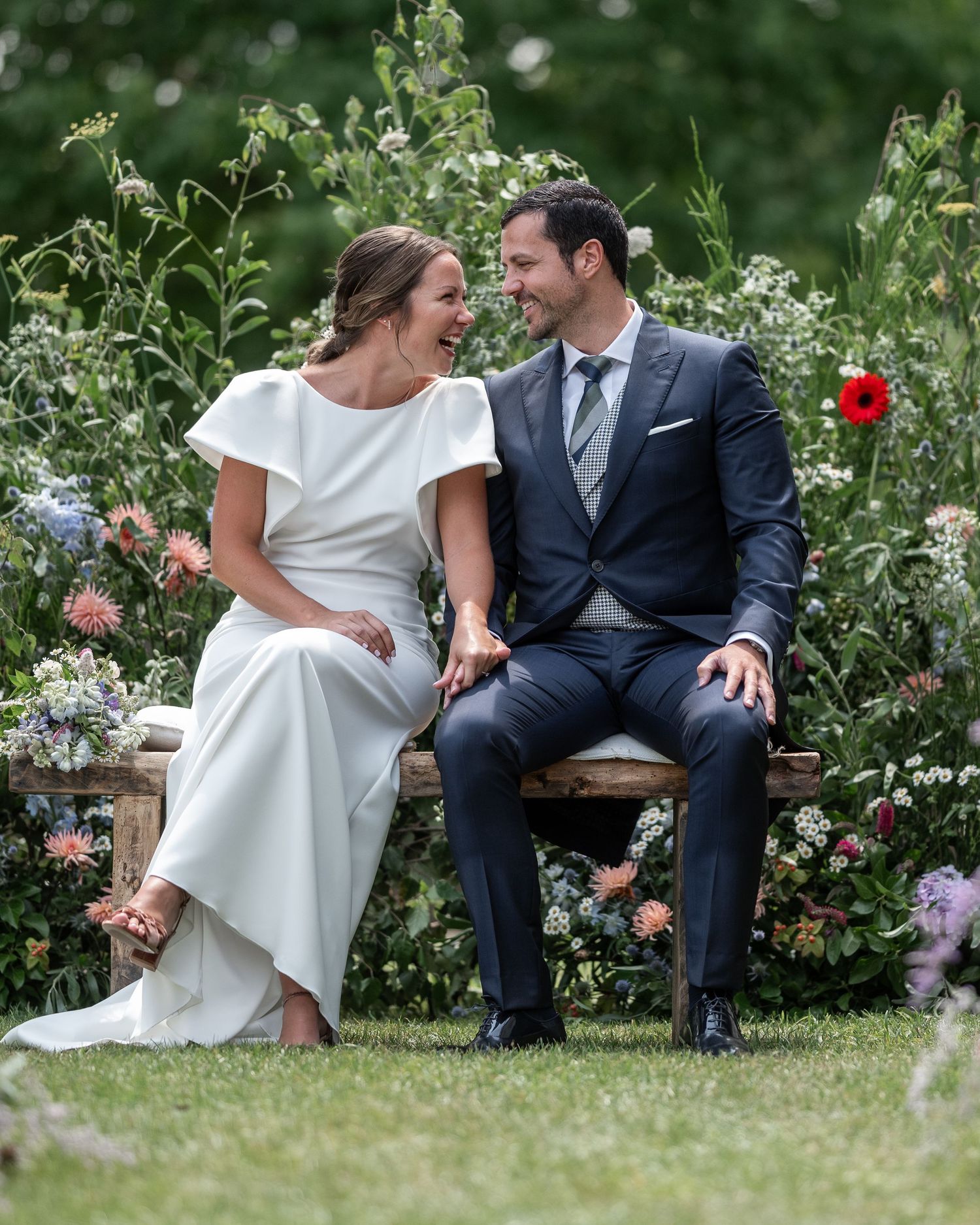 A couple in wedding attire sits together on a bench in a lush garden setting with blooming flowers.