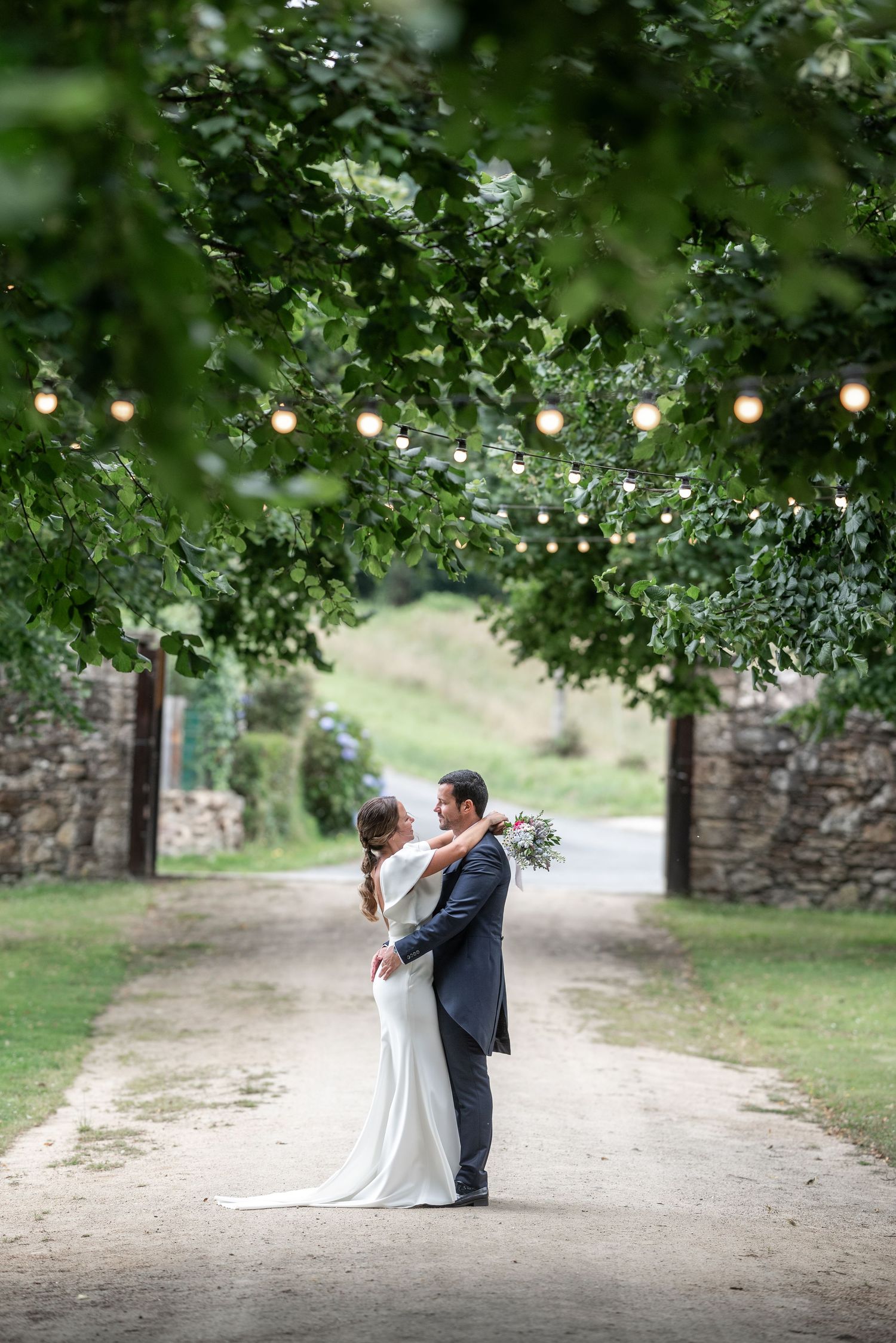 A couple in wedding attire share a romantic moment under a canopy of green trees on a dirt path.