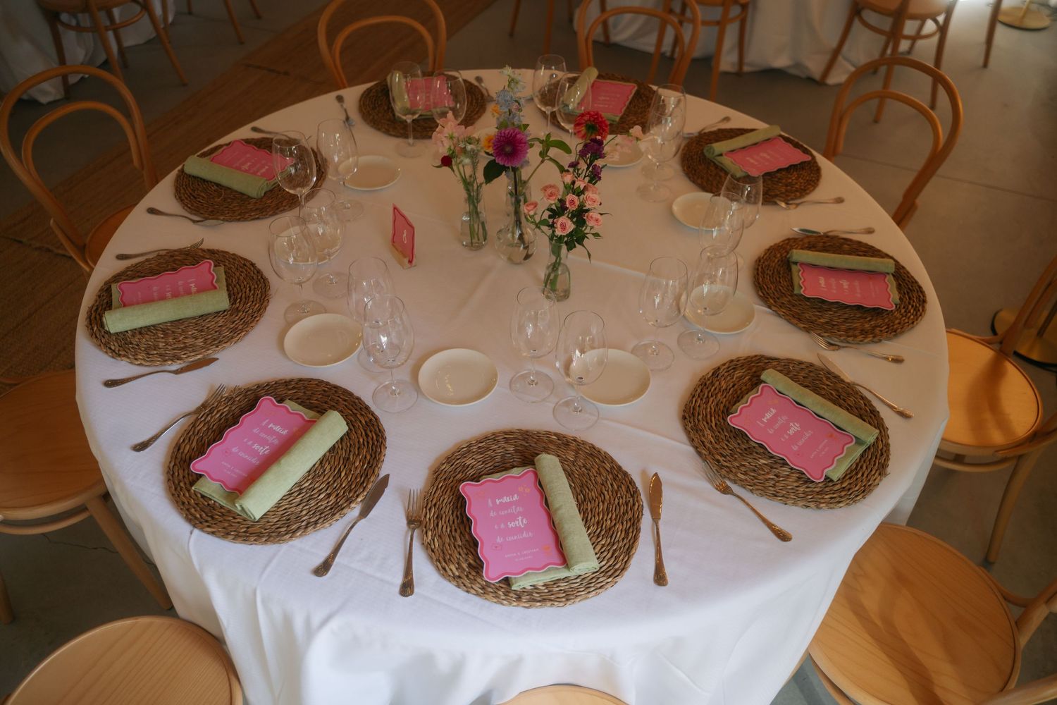A round table set for a wedding reception with white linens, charger plates, pink place cards and a floral centerpiece.
