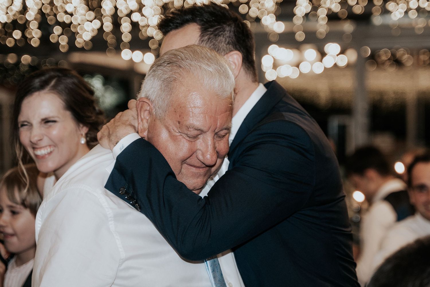 Wedding guests gather at an elegant indoor reception venue illuminated by warm string lights and chandeliers.