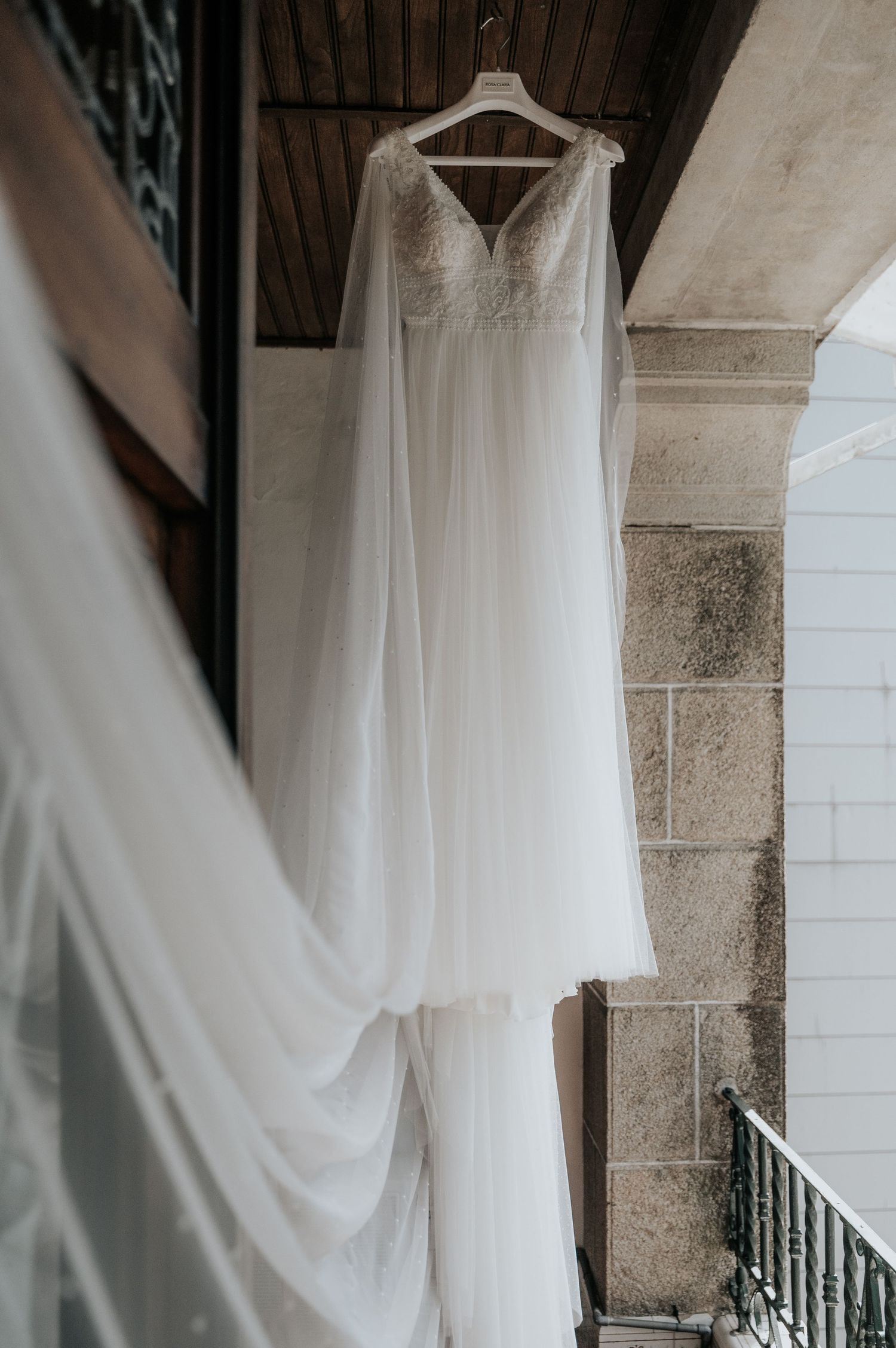Elegant white wedding dress hanging on display against a rustic stone wall and iron railing.