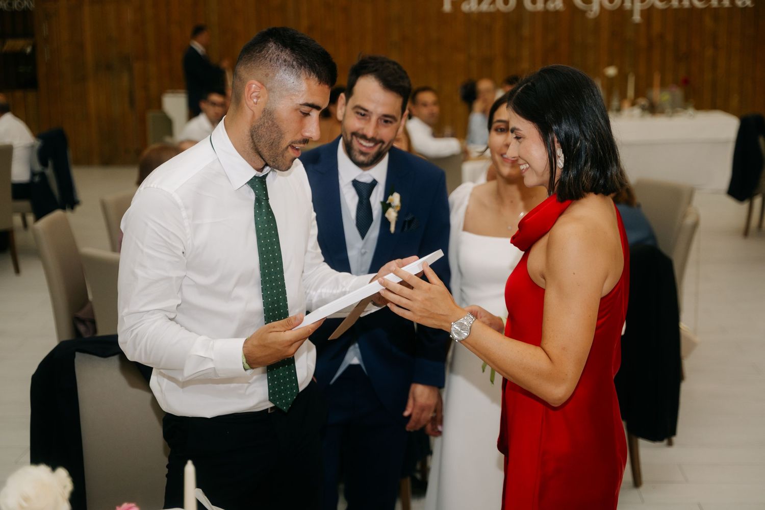 Wedding guests share a lighthearted moment while looking at a phone during an indoor reception celebration.