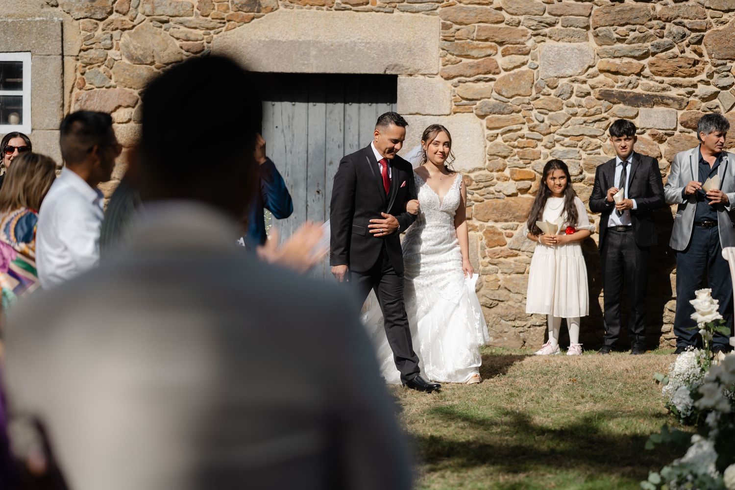 Wedding guests gather outside a rustic stone building during a sunny outdoor celebration.