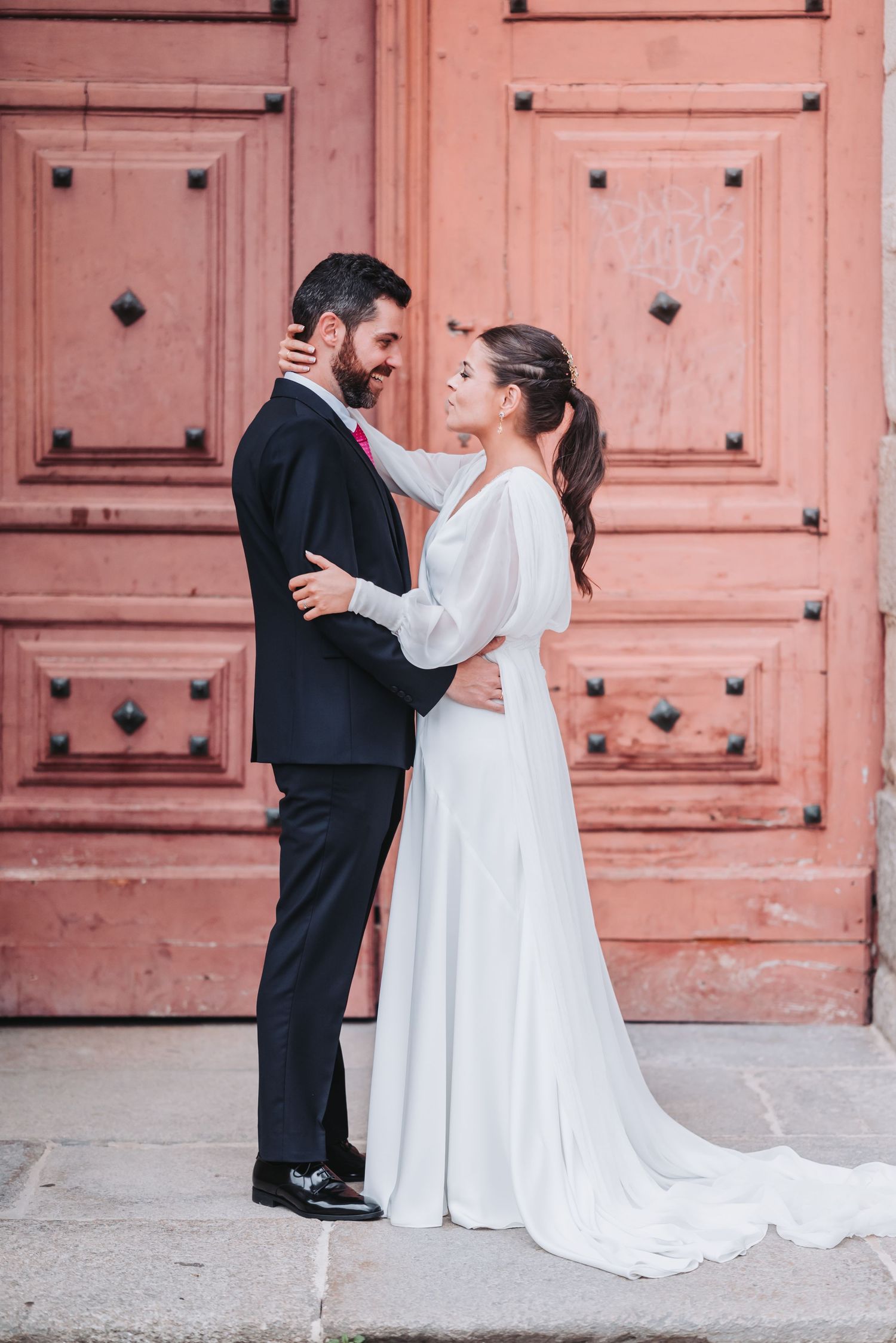 A couple in wedding attire dances together in front of a large pink door in a romantic first dance pose.
