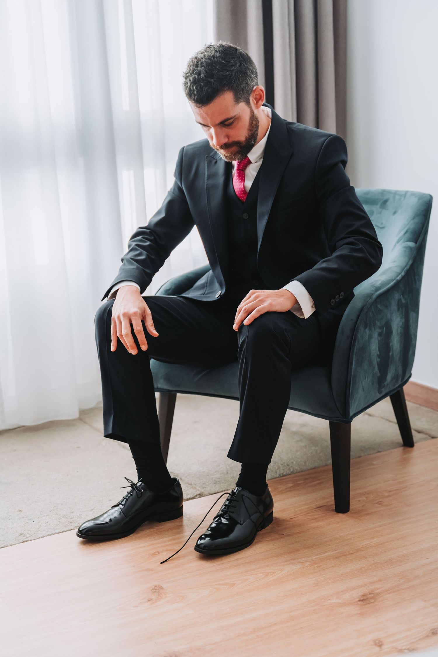 Person in black suit sitting in dark armchair tying dress shoes on wooden floor.