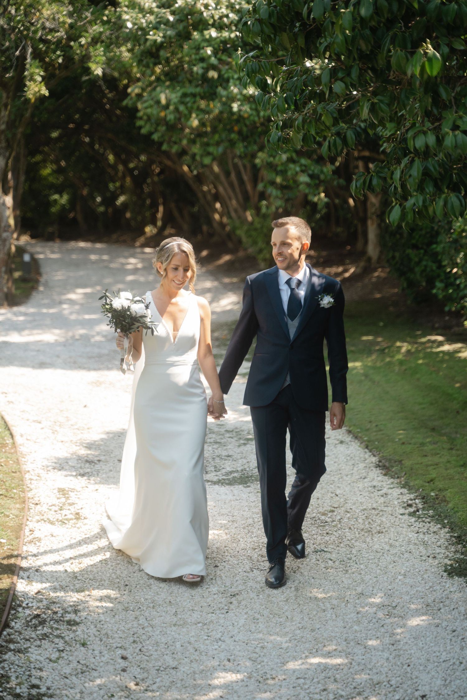 A couple walks hand in hand down a gravel path surrounded by lush greenery on their wedding day.